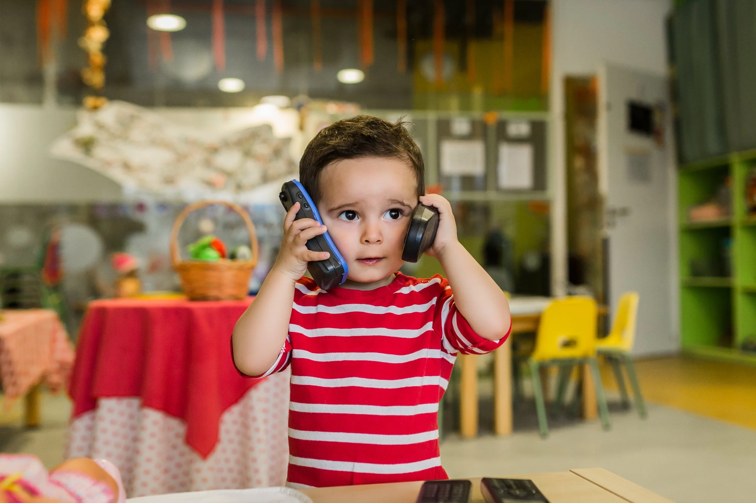 A young child wearing a red and white striped shirt holding two phones to his ears in a colorful room with tables and chairs.