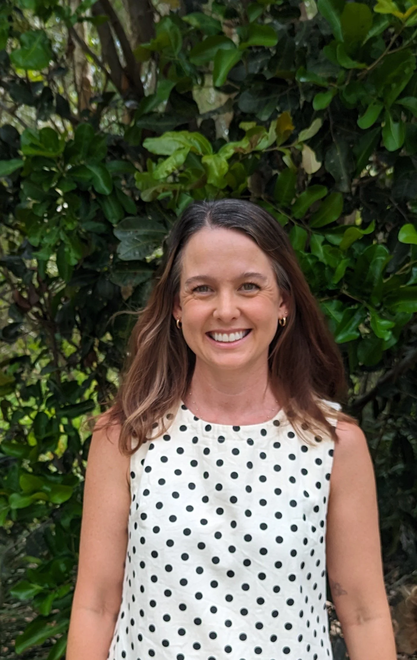 A young woman smiling outdoors, wearing a white sleeveless top with black polka dots, standing in front of green leafy bushes.