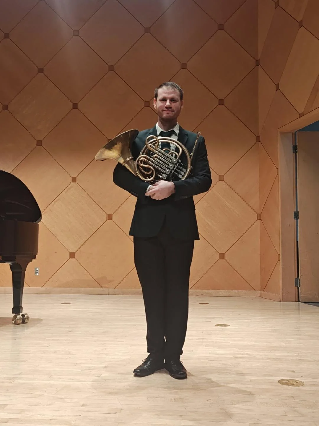 Man in formal black suit holding a French horn standing on stage with wooden wall background and a black grand piano.