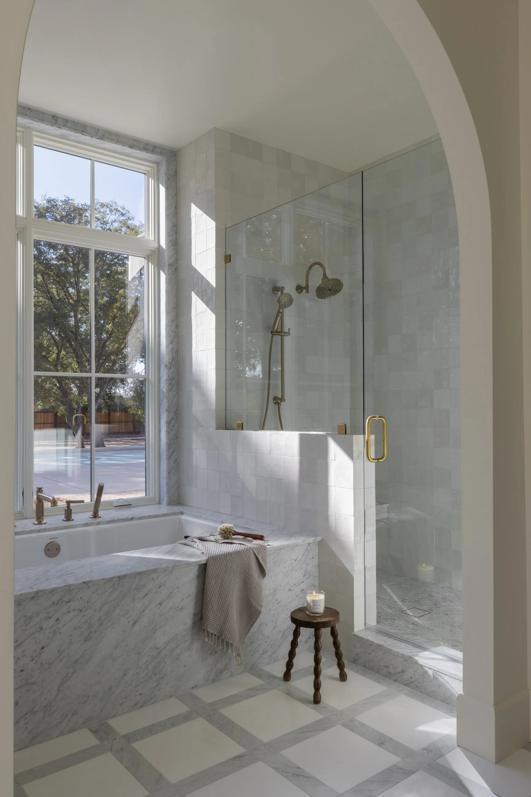 Bright bathroom with a large window, marble bathtub, glass-enclosed shower, candle, and a small wooden stool.