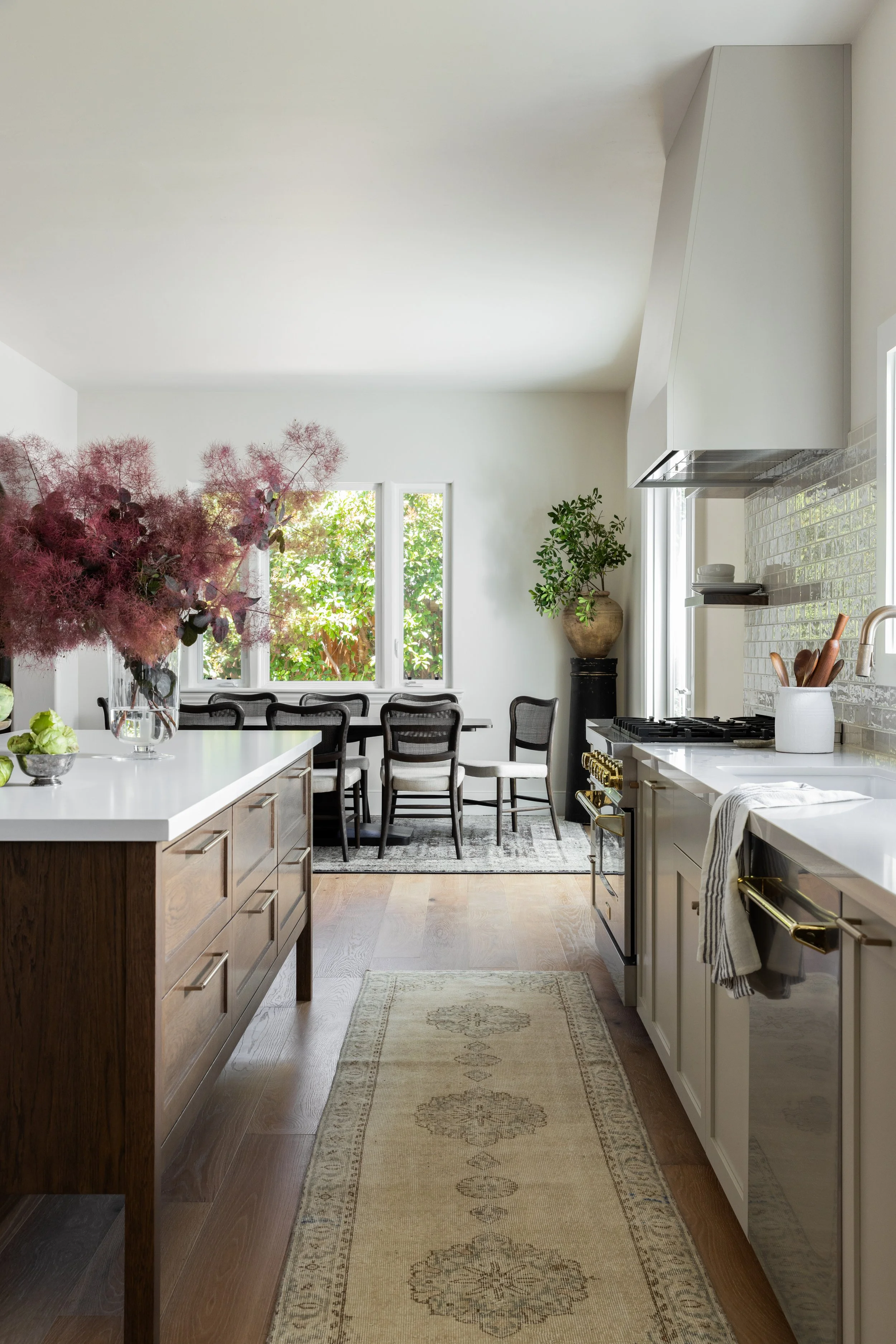 Modern kitchen with a wooden island, white countertops, a gas stove, and a dining area with dark chairs, a window, plants, and natural light.