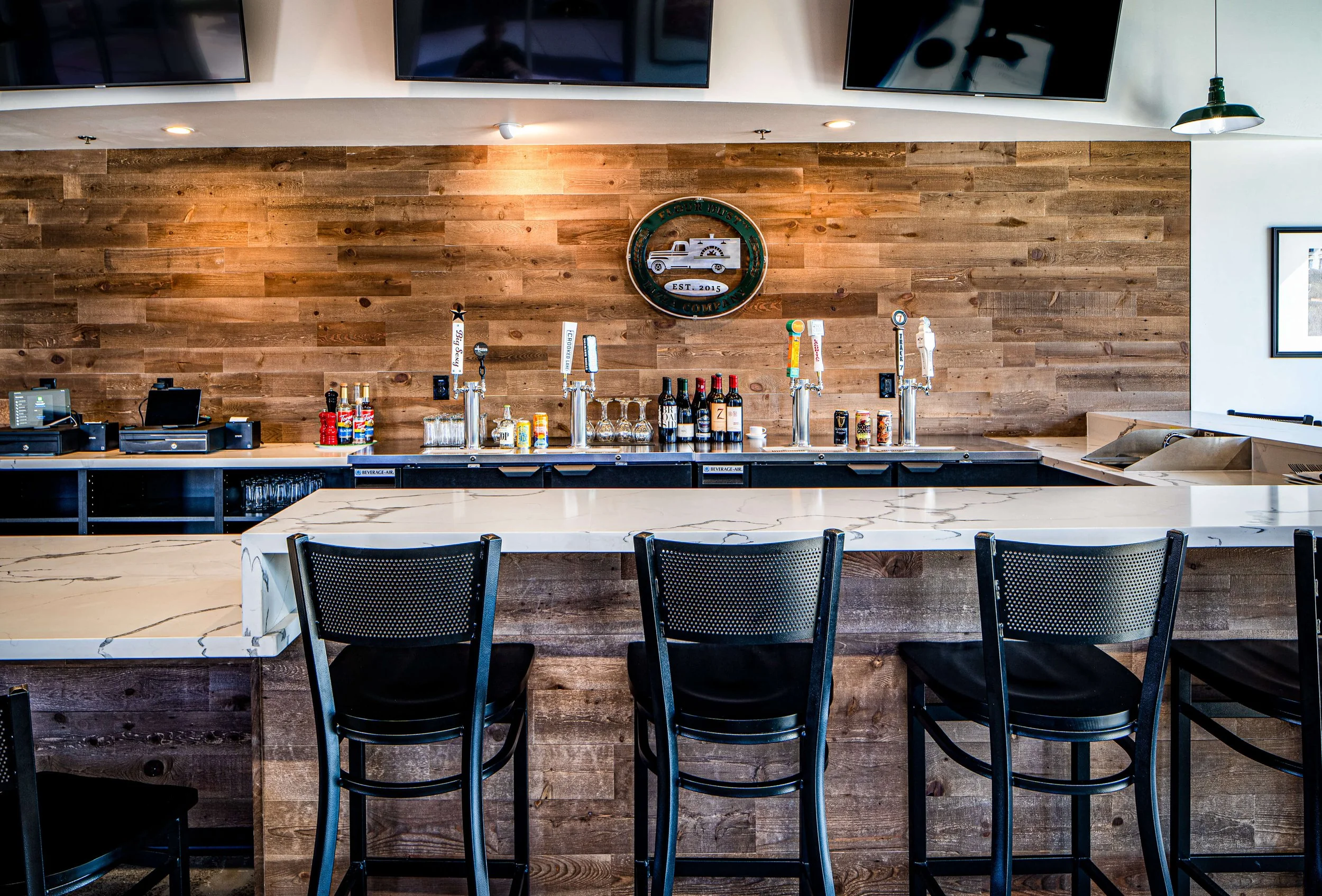 Sacramento CA commercial renovation and design with an empty bar with black chairs, taps, bottles, glasses, and a wood-paneled wall with a logo.