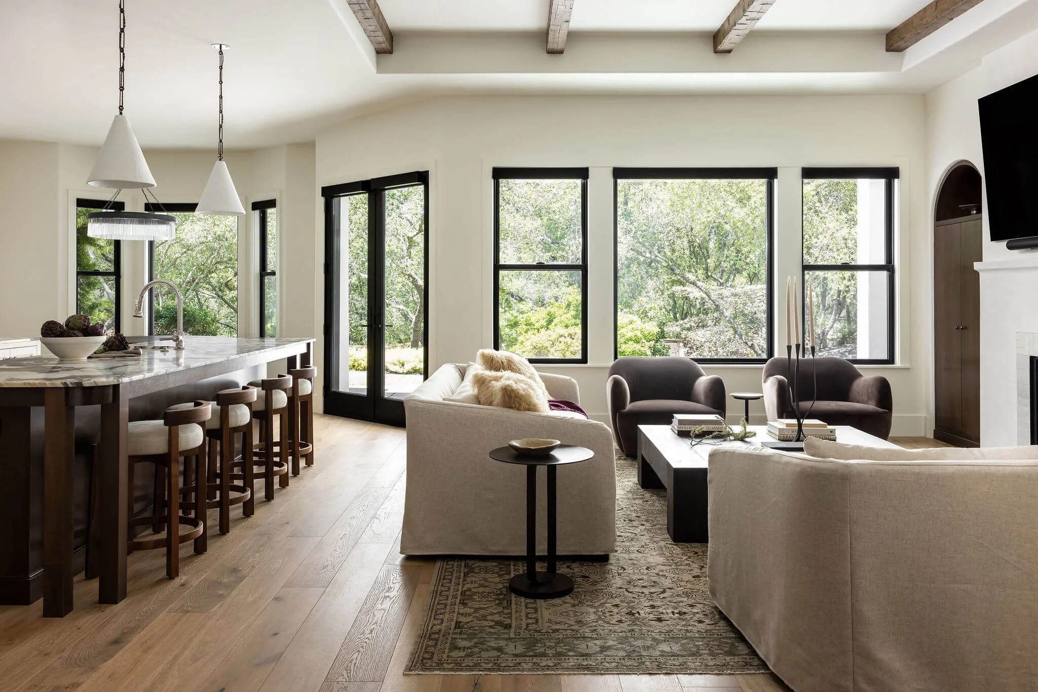 Bright living room with large windows, beige sofas, dark armchairs, a black coffee table, a rug, and a view of trees outside.