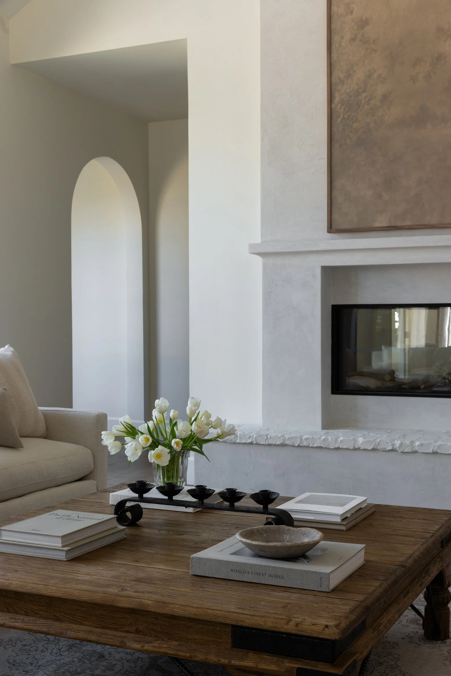 Custom home with refined living room with beige sofa, wooden coffee table, white flowers in a glass vase, books, a stone bowl, fireplace, and minimalist decor.