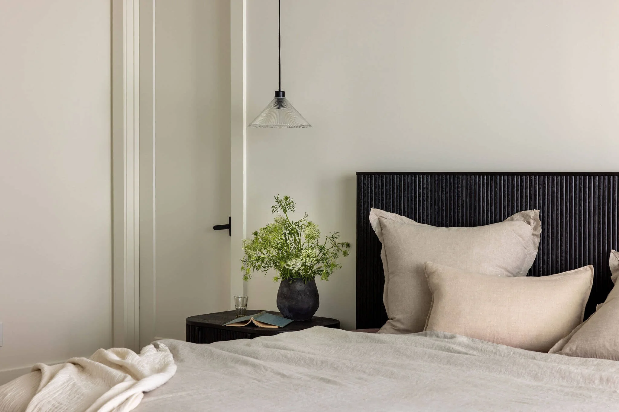 A bed with black headboard, a black nightstand with a green plant and book, and a simple pendant light