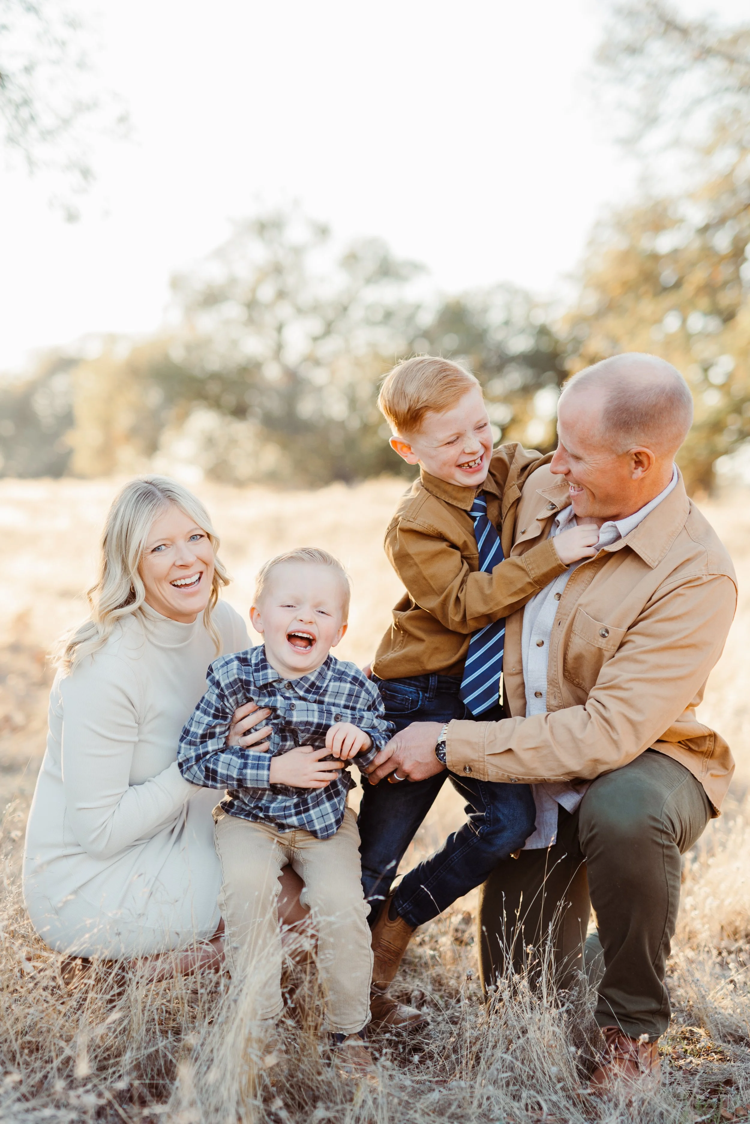 Family of four laughing and playing outdoors in a grassy field with trees in the background during daytime.