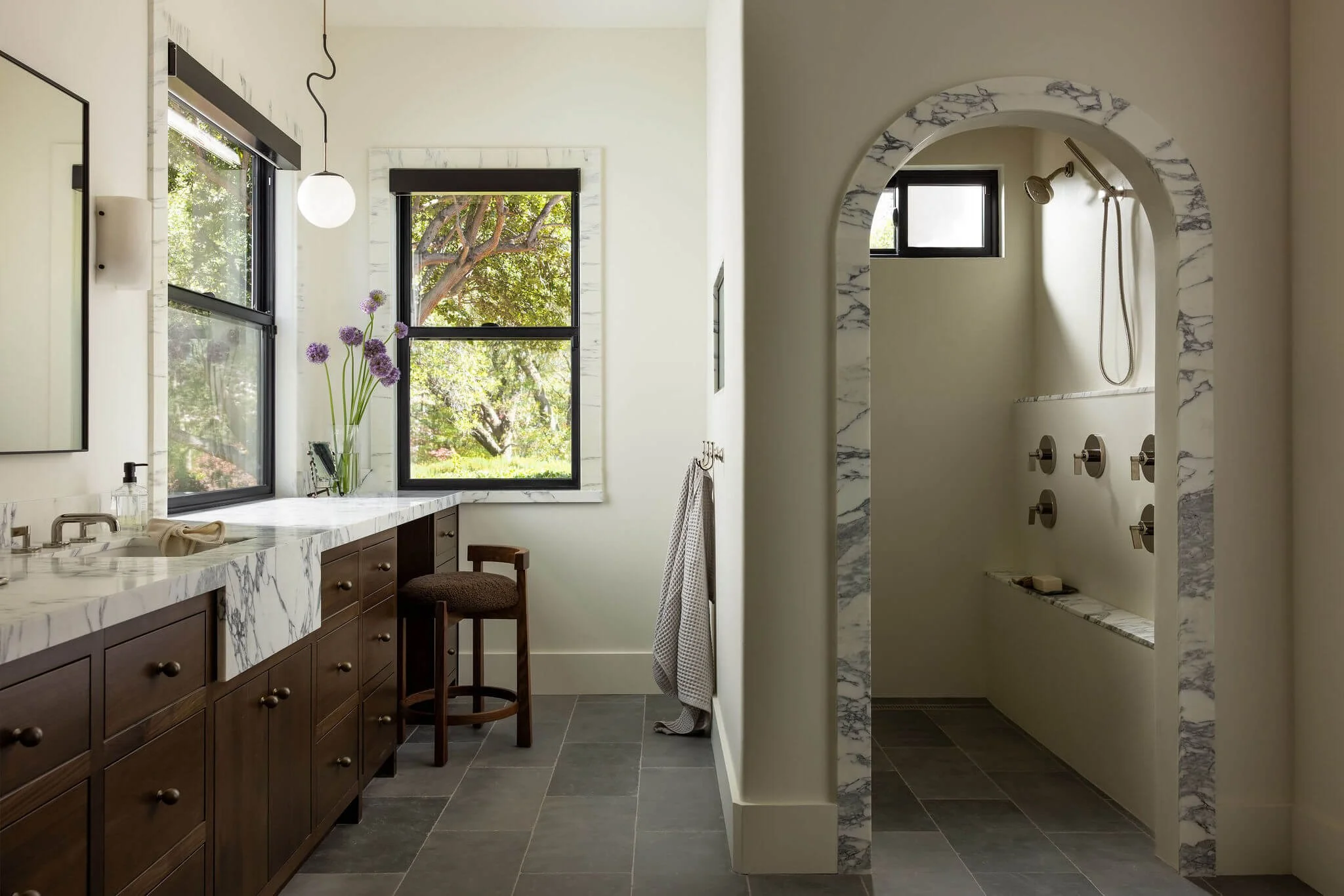 A bathroom with a marble countertop, dark wooden cabinets, and two black-framed windows overlooking a green, leafy outdoor scene located in Sacramento Home Renovations and additions