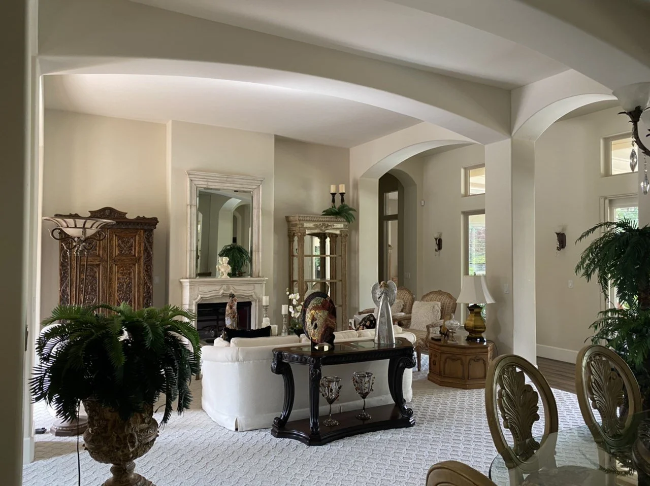 Elegant living room with cream-colored sofas, ornate wooden furniture, tall plants, and decorative items on tables and shelves, bright with natural light from windows.