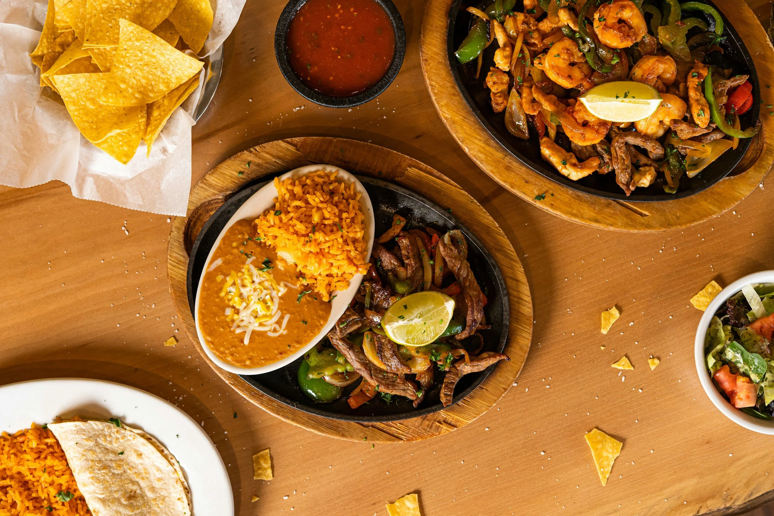 Mexican cuisine with nachos, rice, beef and shrimp fajitas, salsa, and salad on a wooden table.