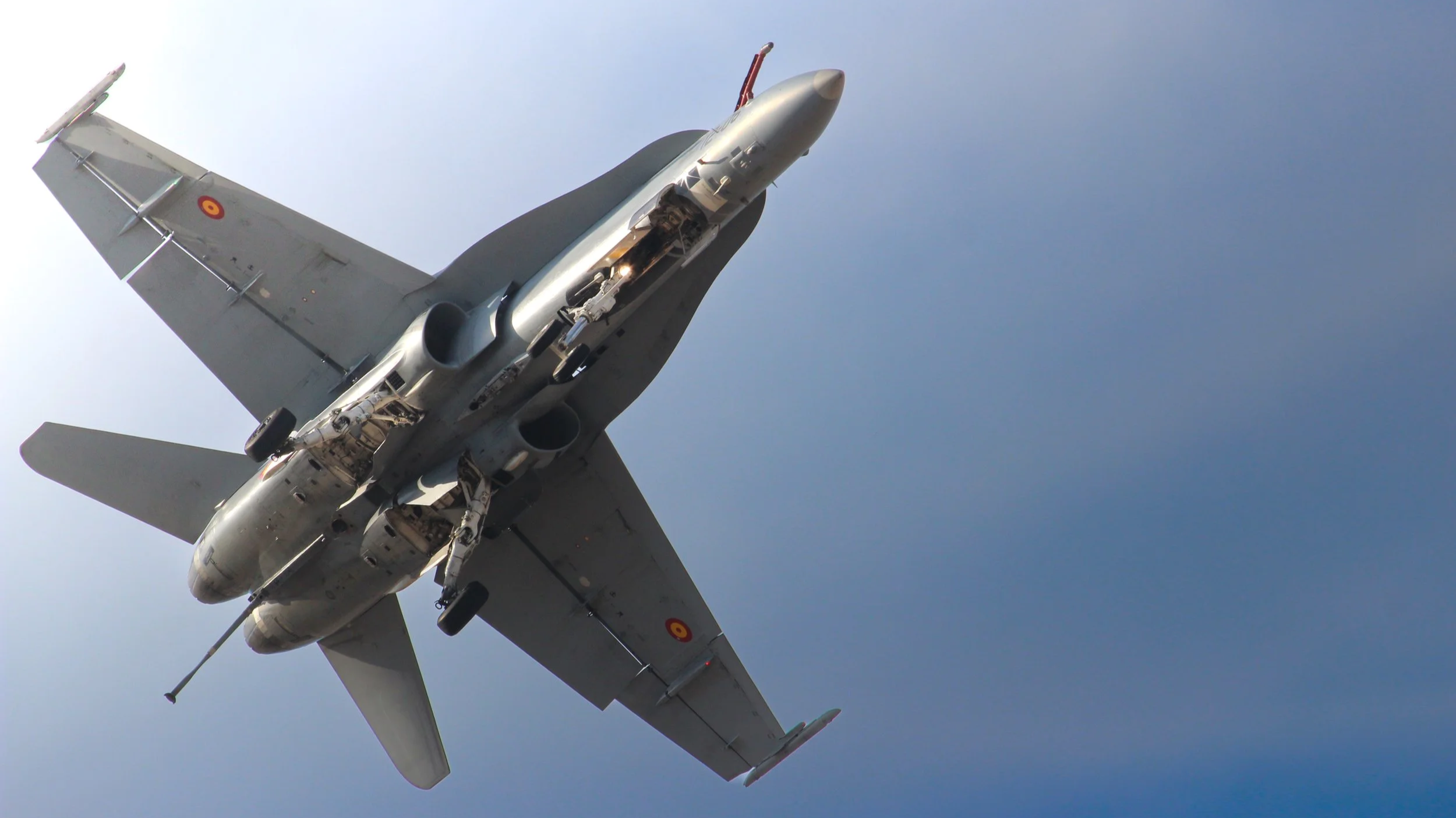 A military fighter jet flying in the sky with the underside visible, showing its wings, engines, and landing gear.