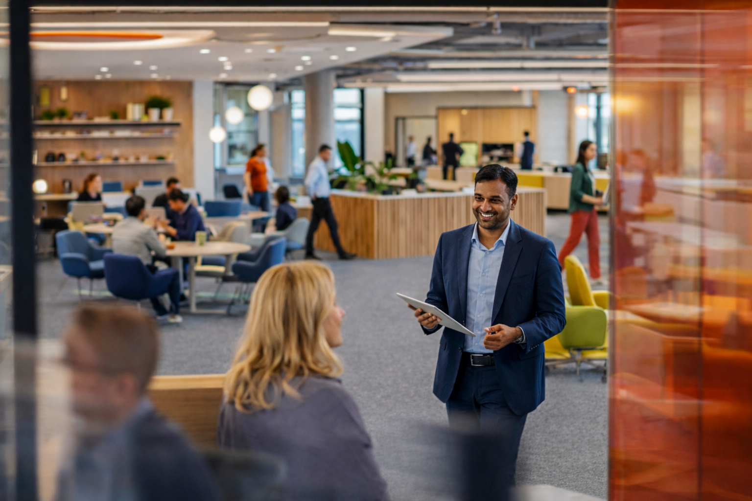 Smiling man in a blue suit holding a tablet, standing and talking to woman with blonde hair in a busy modern office lounge with people working and walking in the background.