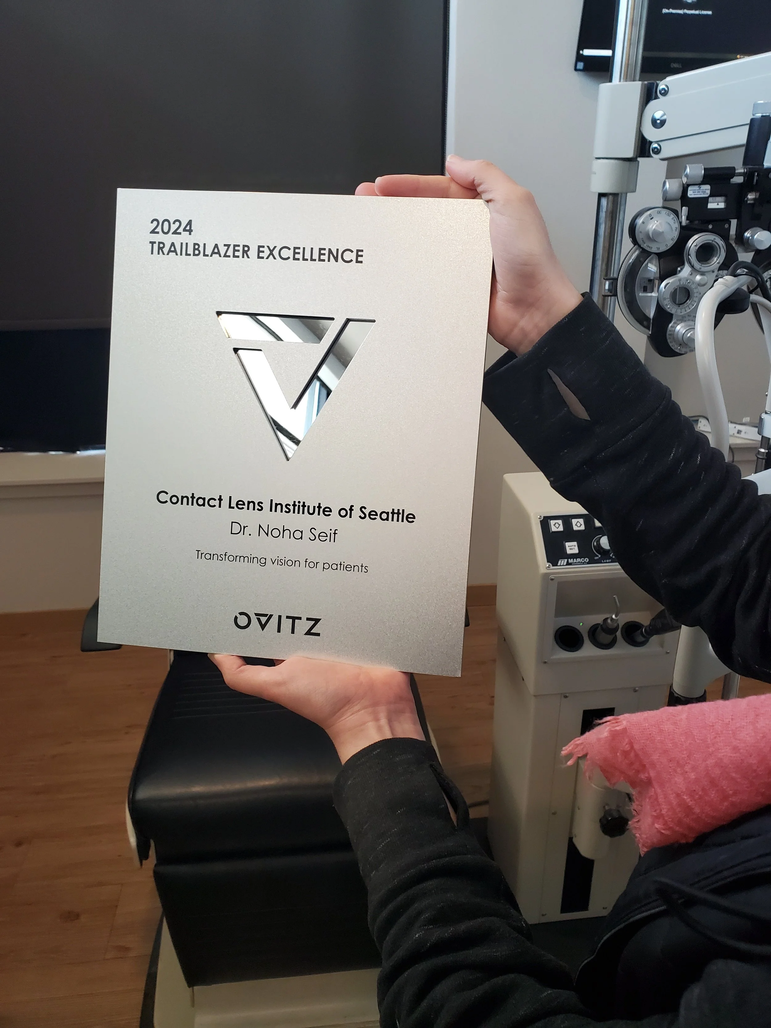 Person holding a certificate from the Contact Lens Institute of Seattle for Dr. Noha Seif, awarded for 2024 Trailblazer Excellence, in a clinical setting with medical equipment in the background.