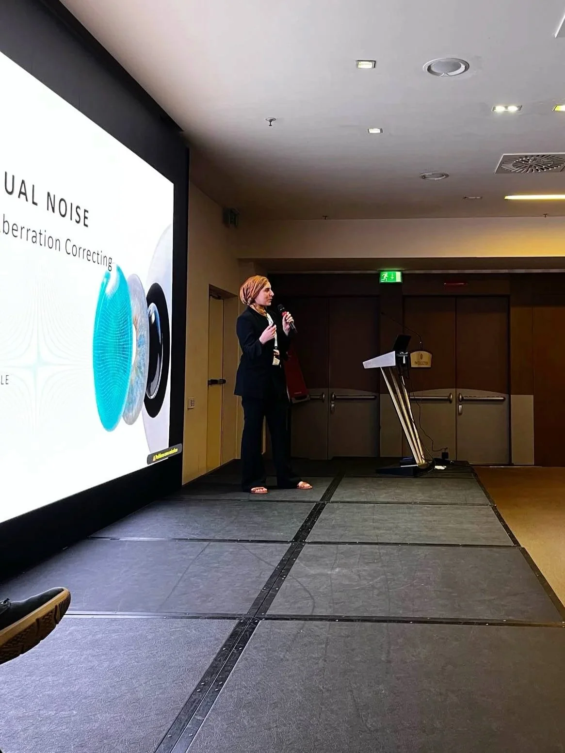 Woman in black suit giving a presentation on a stage with a large screen showing a slide about noise cancellation technology.