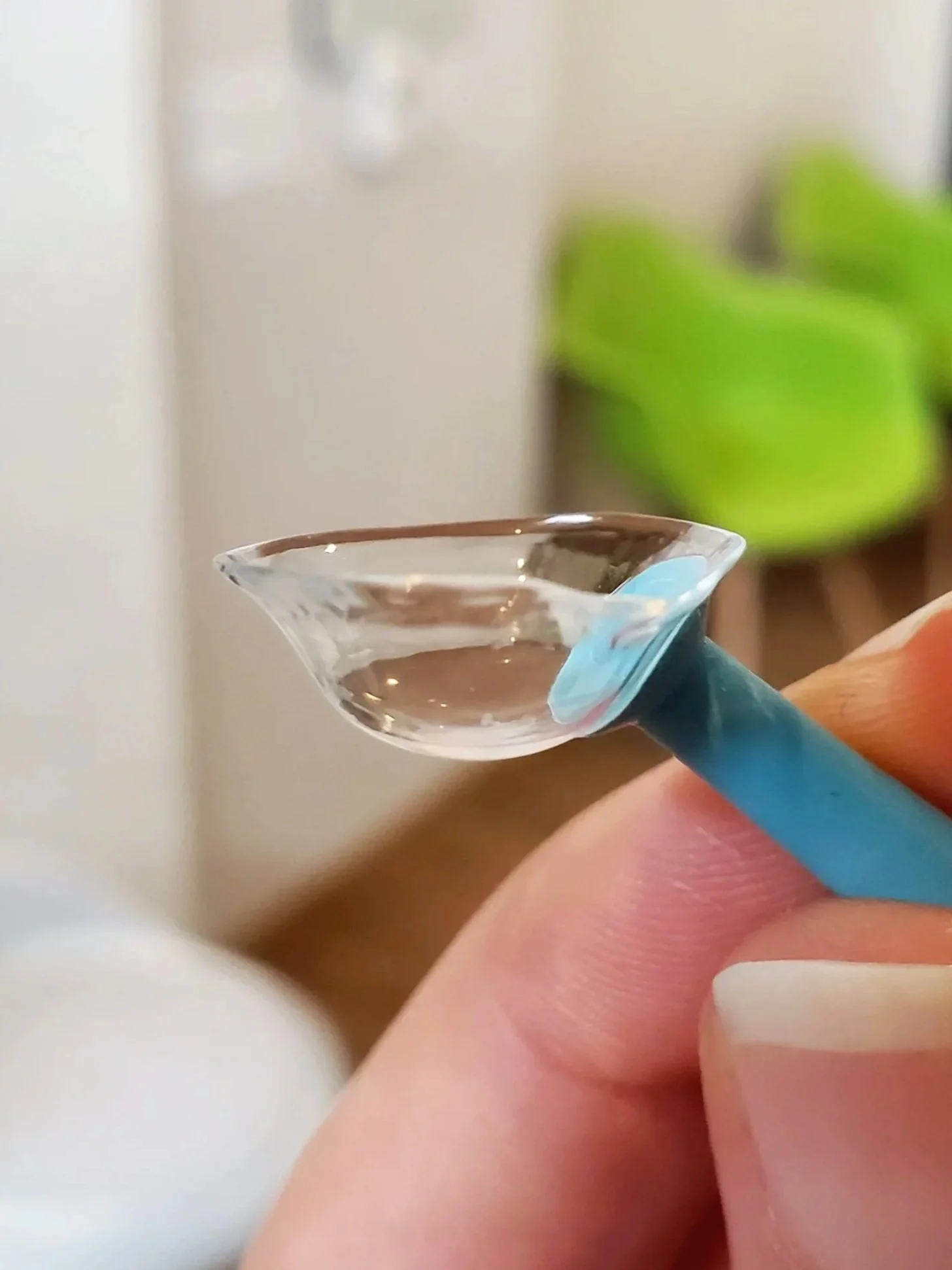 Close-up of a person's hand holding a small, clear glass pipette filled with a transparent liquid, with a blurred indoor background.