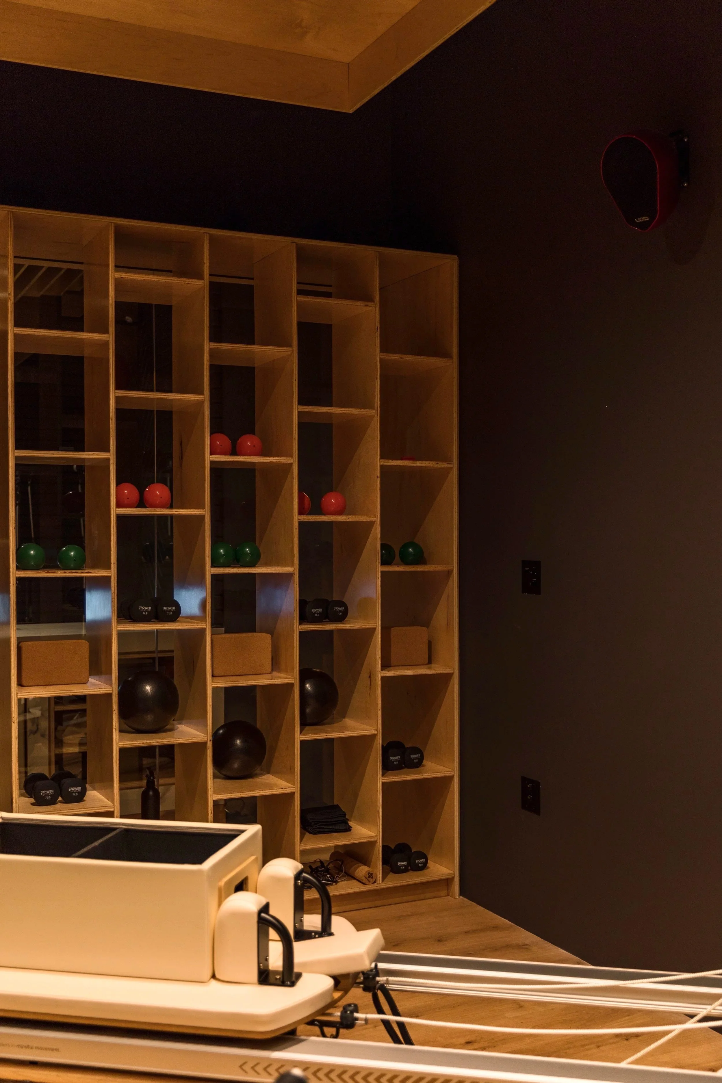 A fitness or yoga room with a wooden shelving unit holding exercise balls, mats, and dumbbells. There is a mirrored wall and a black wall-mounted speaker in the corner.