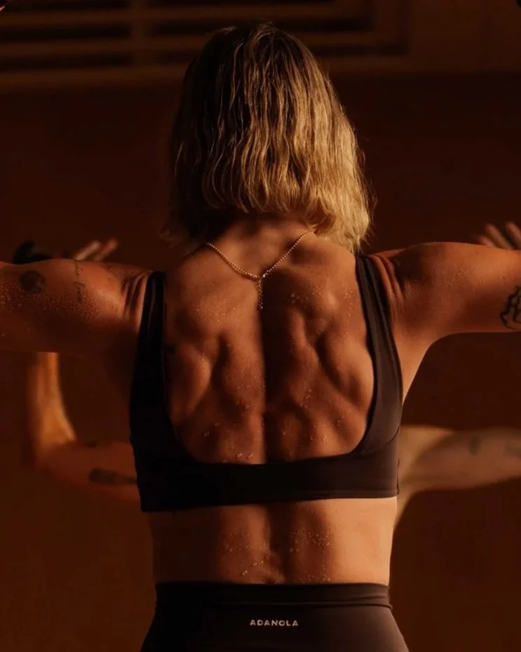 A woman with short blonde hair seen from the back, wearing a black sports bra and black leggings, flexing her arms in a gym or fitness studio.