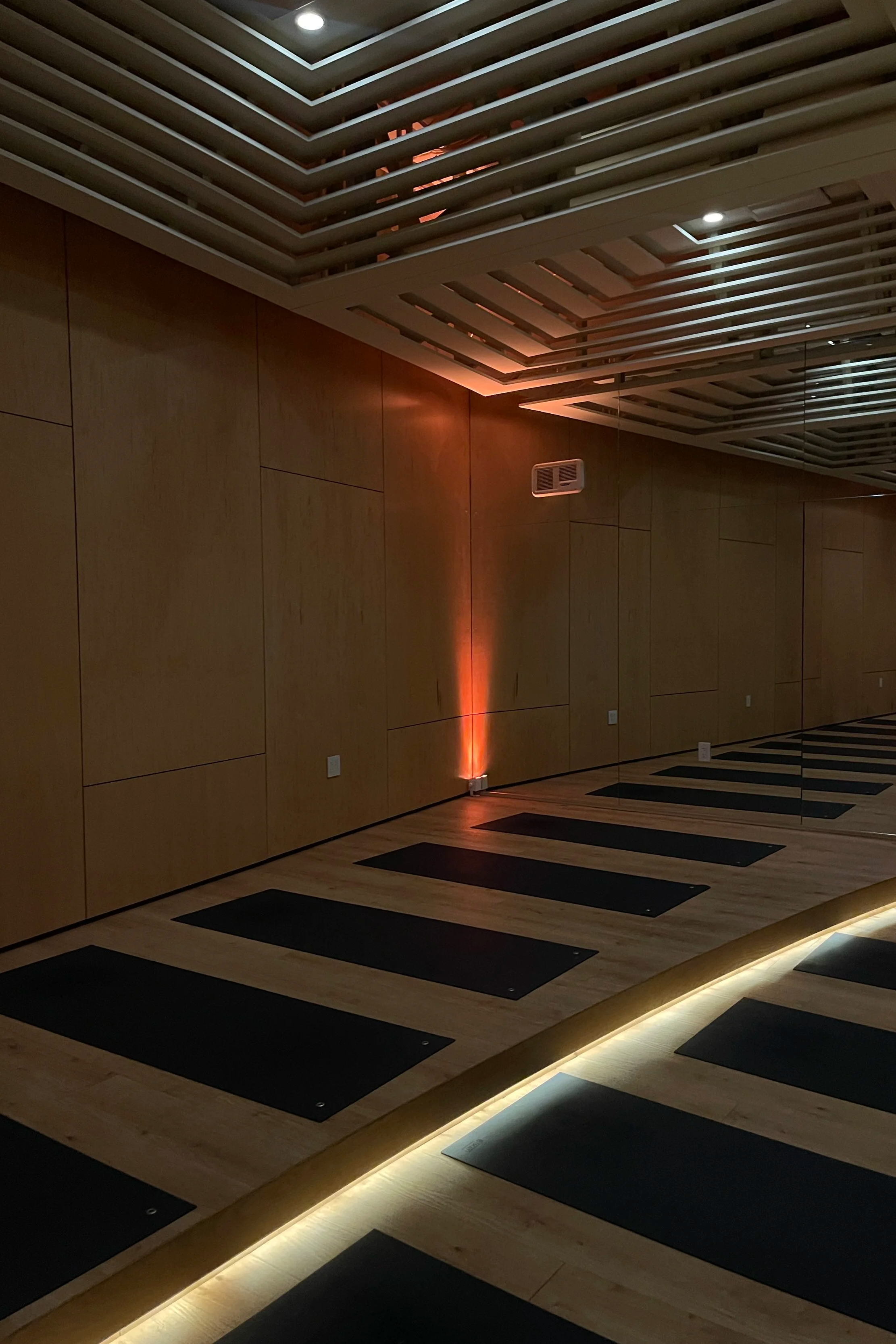 Interior of a yoga or exercise studio with black mats arranged on a wooden floor, illuminated by soft orange and white lighting, with wood-paneled walls and a ceiling featuring white slats and recessed lights.