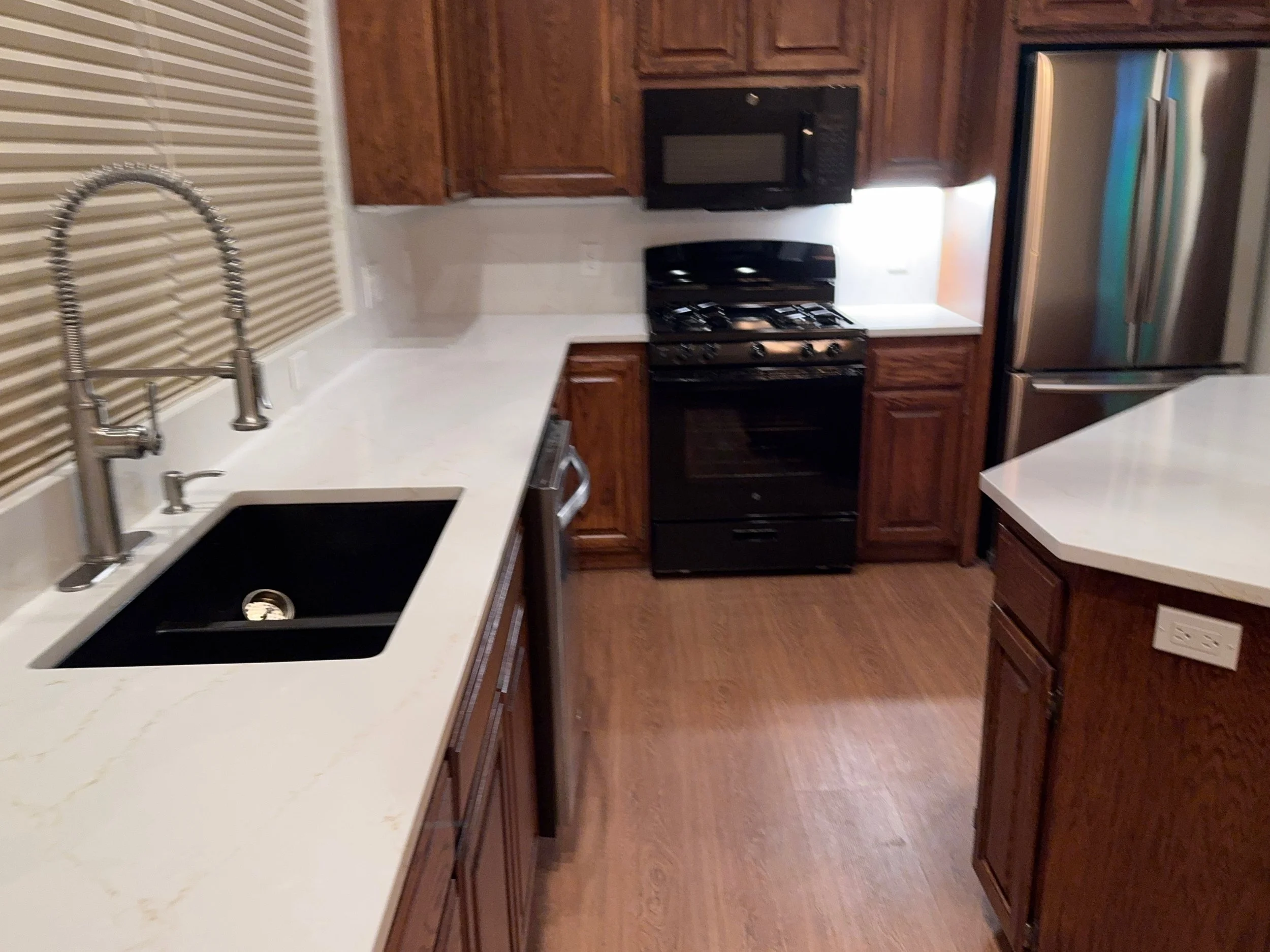 A kitchen with wooden cabinets, white countertops, a black sink, a stainless steel refrigerator, a black stove, a microwave, and a window with beige blinds.