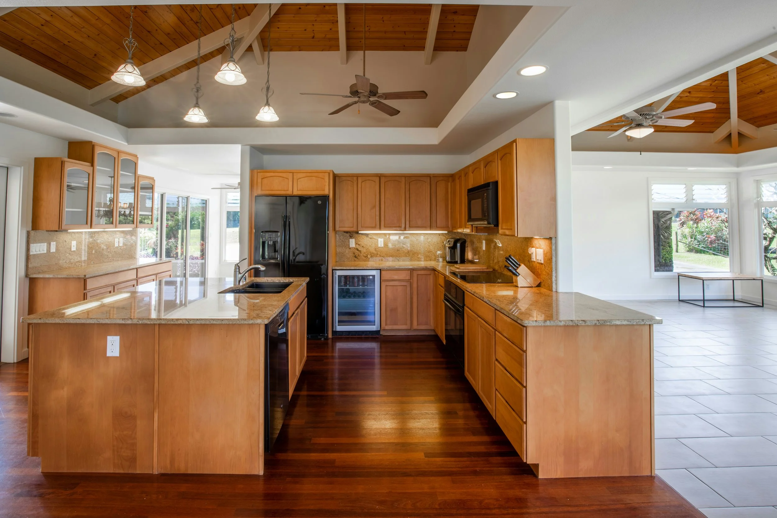 Modern kitchen with wooden cabinets, granite countertops, black appliances, and a view of a bright living space with large windows and a tiled floor.