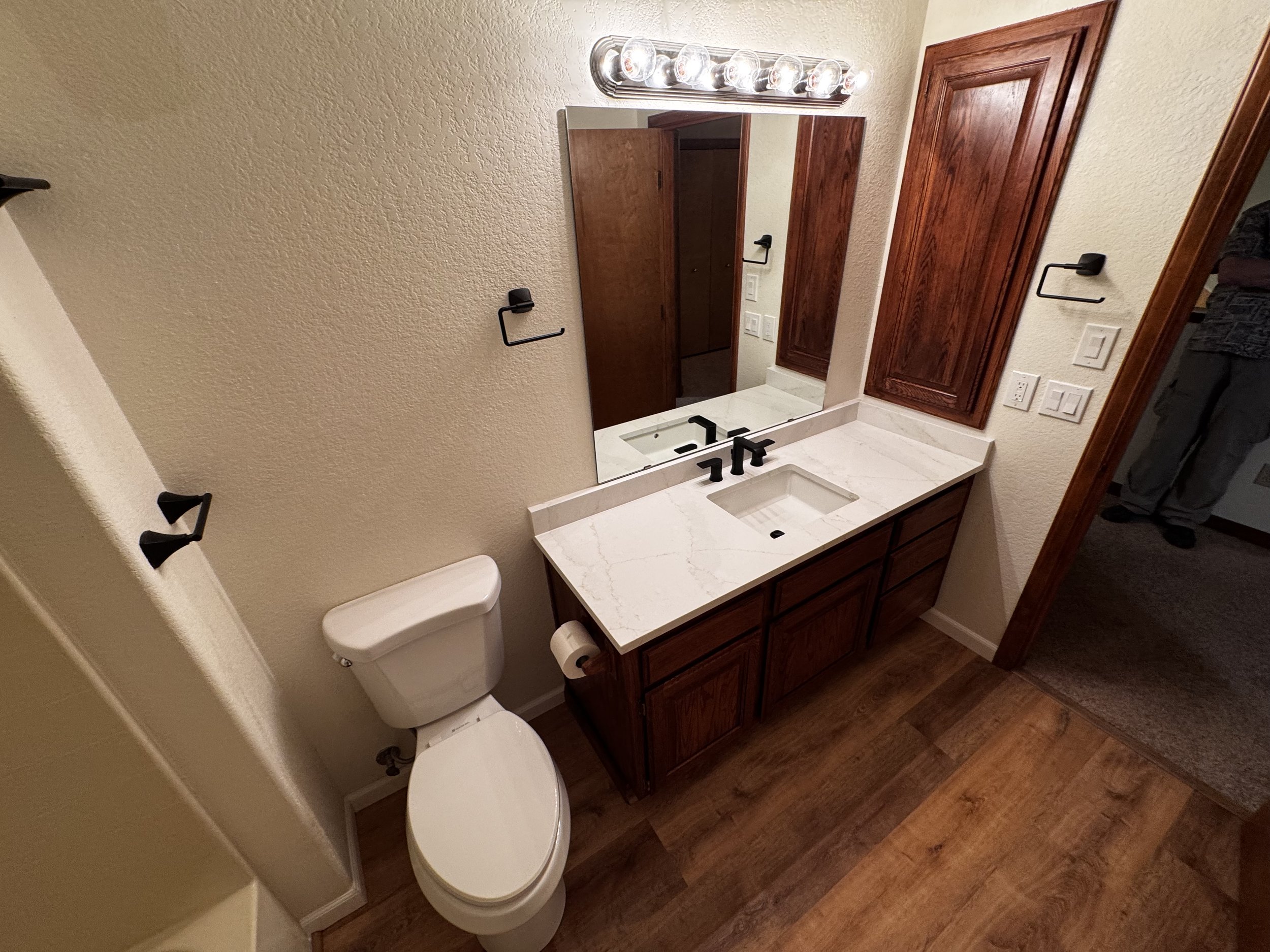 A bathroom with a white toilet and a wooden vanity with a marble countertop, a rectangular sink, black faucet, large mirror, wooden cabinet, and wood floor, with black towel holders and electrical outlets.