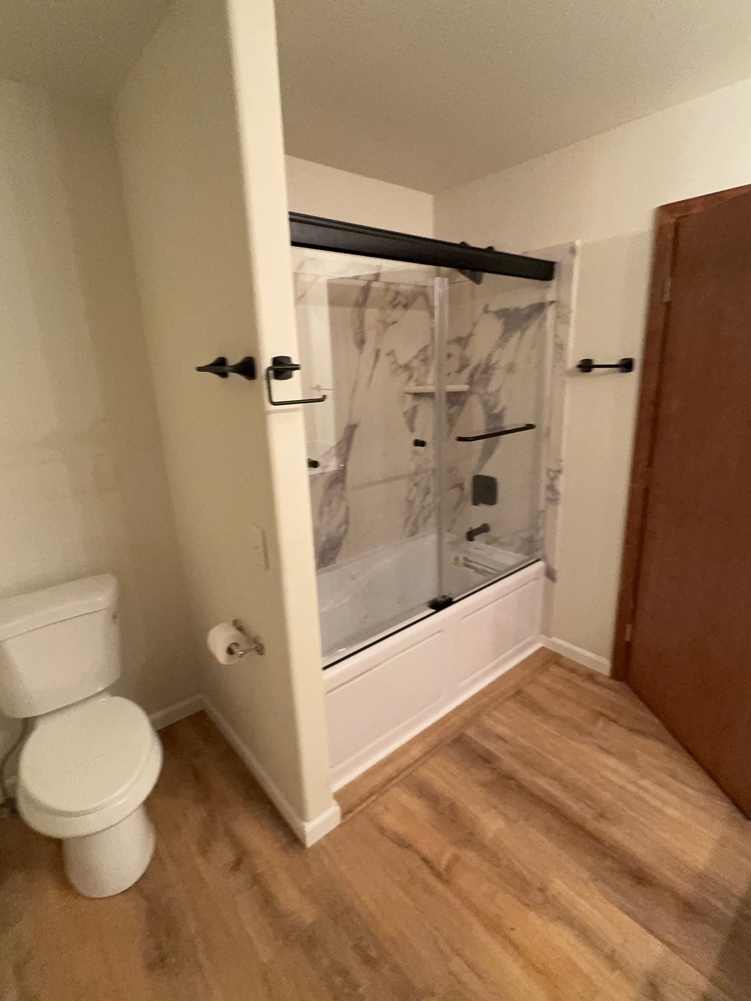 A bathroom with a white toilet on the left, next to a shower stall with a clear glass door and marble-patterned walls. There are black hooks on either side of the shower and a black rod at the top. The floor is wooden.