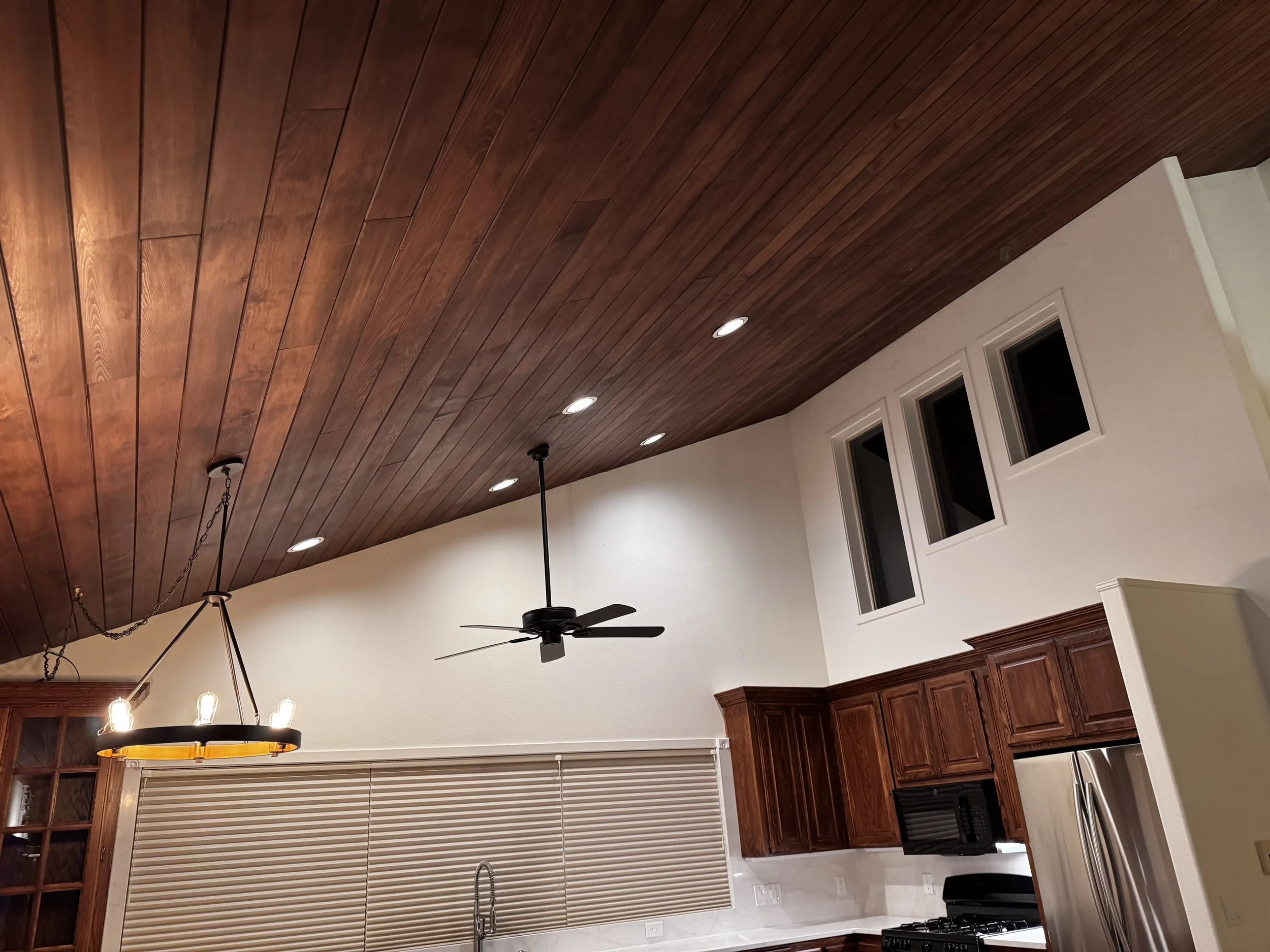 Interior view of a kitchen with a wooden ceiling, ceiling fan, recessed lighting, wooden cabinets, a window with blinds, and a black chandelier with three lights.