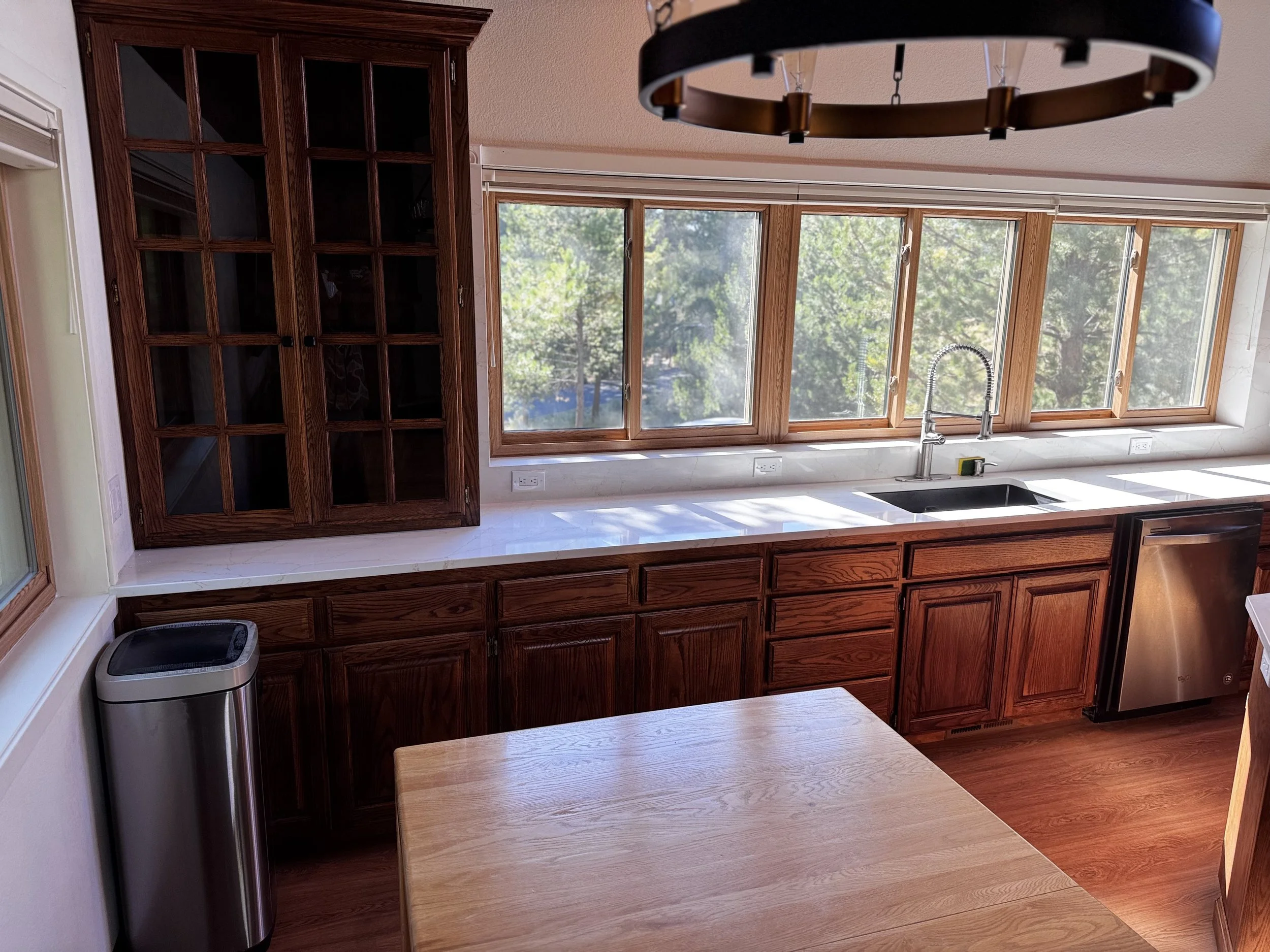 A kitchen with large windows overlooking trees, wooden cabinets, white countertops, a stainless steel trash bin, a black dishwasher, and a modern black and brass circular chandelier.
