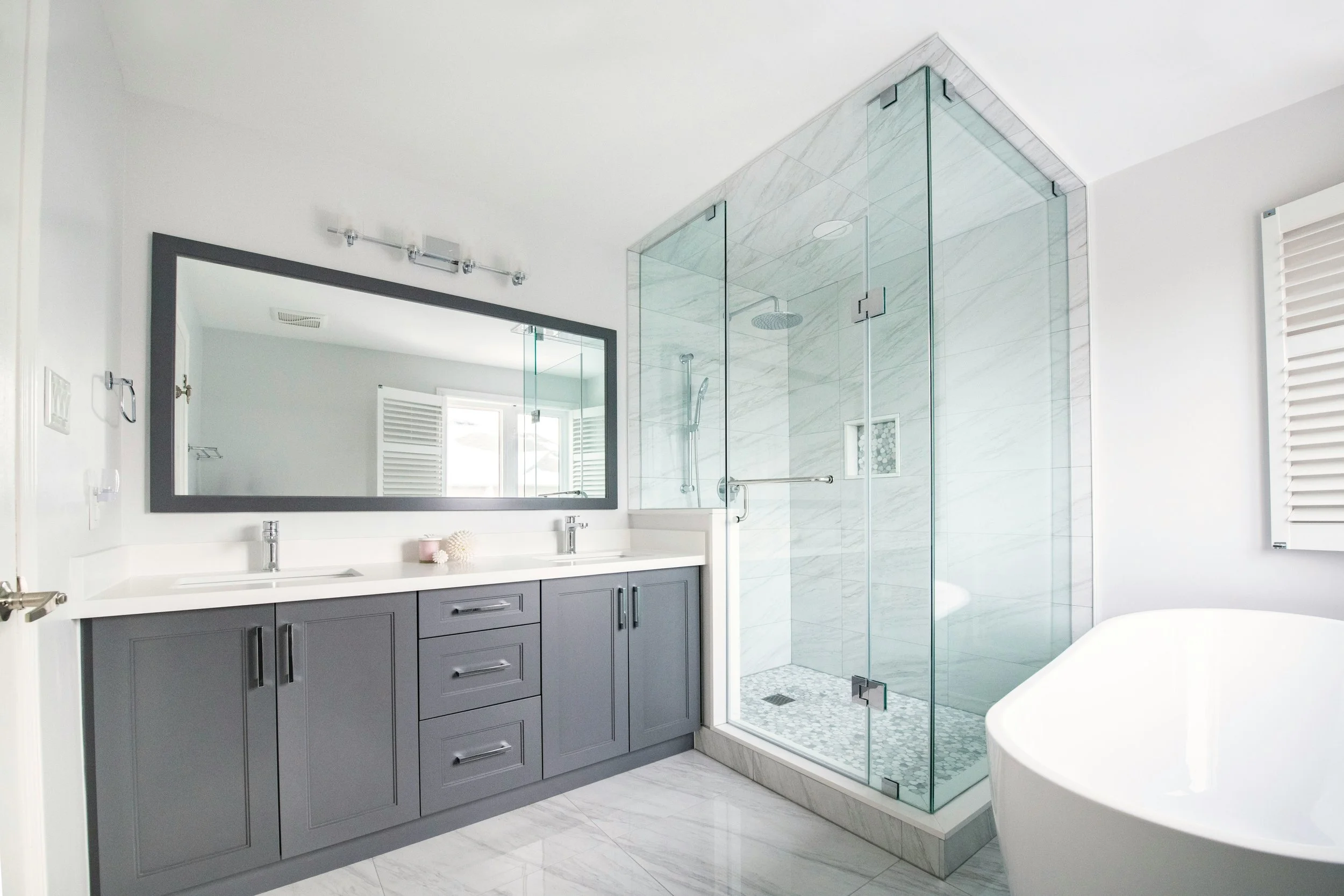 Modern bathroom with gray cabinet vanity, large mirror, glass-enclosed shower with marble tiles, and a white free-standing bathtub.