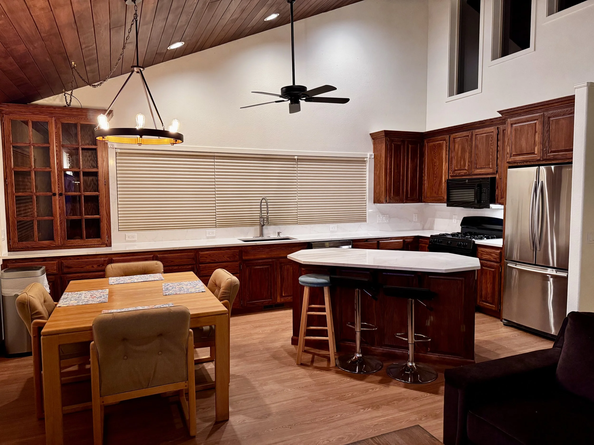 A spacious kitchen with wooden cabinets, a stainless steel refrigerator, a black microwave, a black stove, a white marble countertop with a small breakfast bar and two stools, a sink under a window with blinds, a dining table with four cushioned chairs, a ceiling fan, a unique chandelier, and a high vaulted ceiling with recessed lights.
