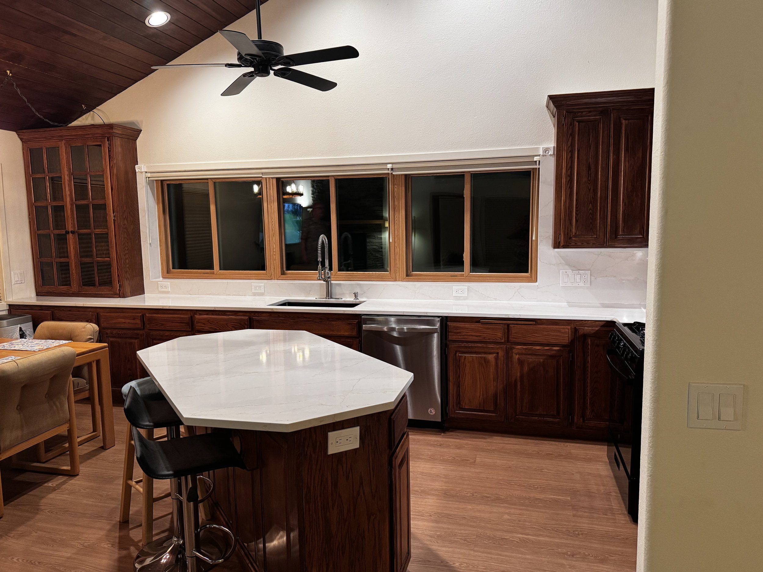 Kitchen with wooden cabinets, a white marble countertop, a kitchen island, a stainless steel dishwasher, a black stove, a large window behind the sink, and a ceiling fan.