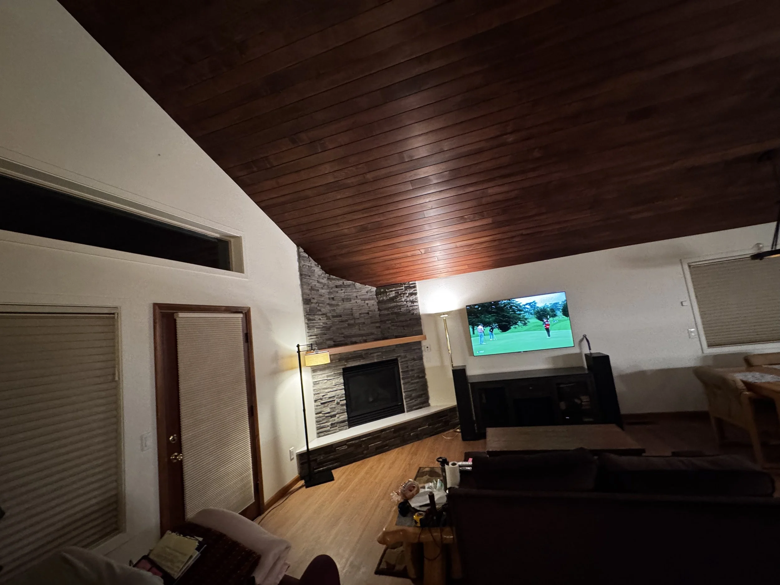 Living room with a wooden ceiling, stone fireplace, television showing a golf game, and furniture including a sofa and chairs.
