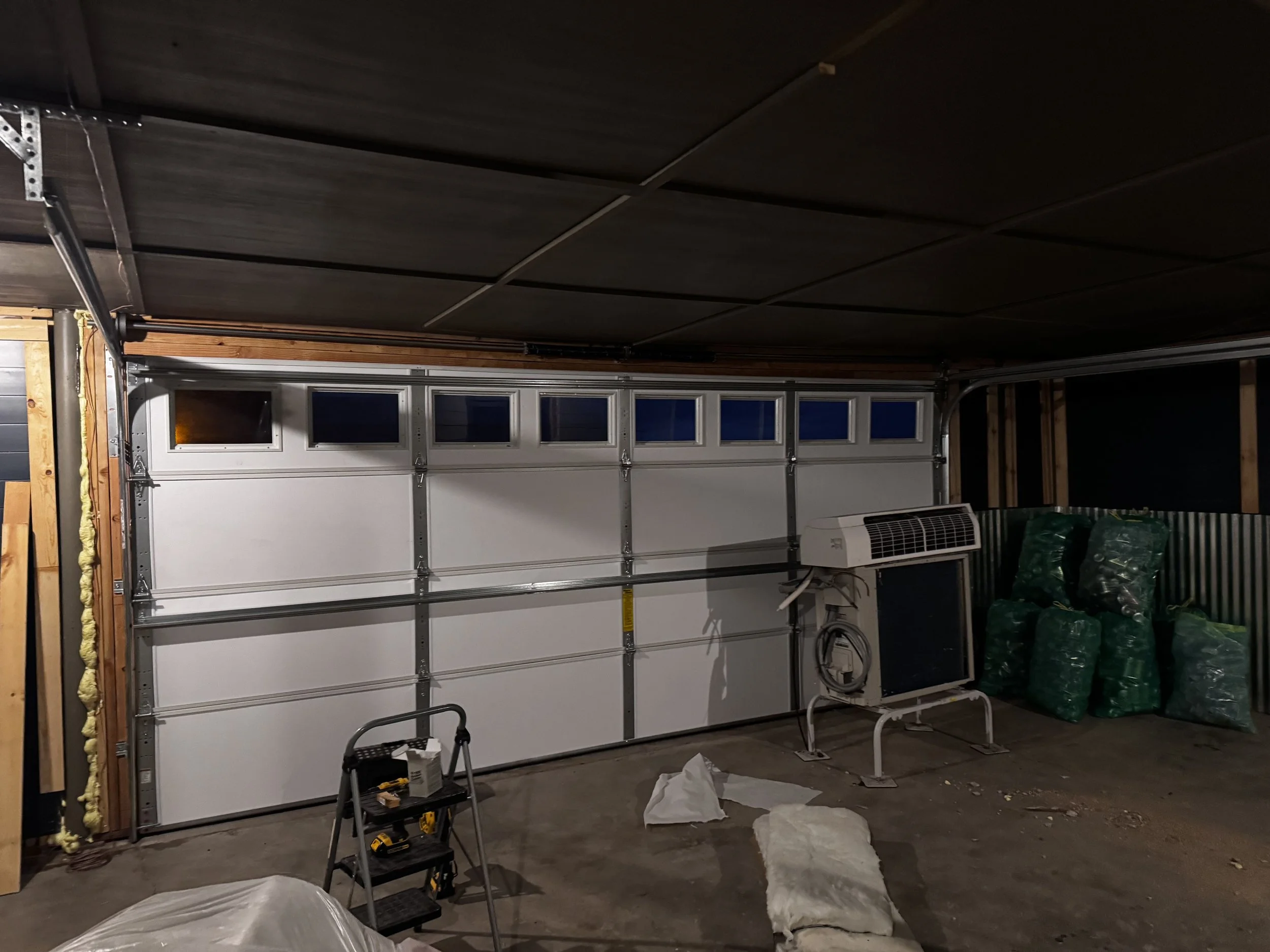 Garage with a white roll-up door, a window air conditioning unit, and bags of green plastic bottles stacked in one corner.