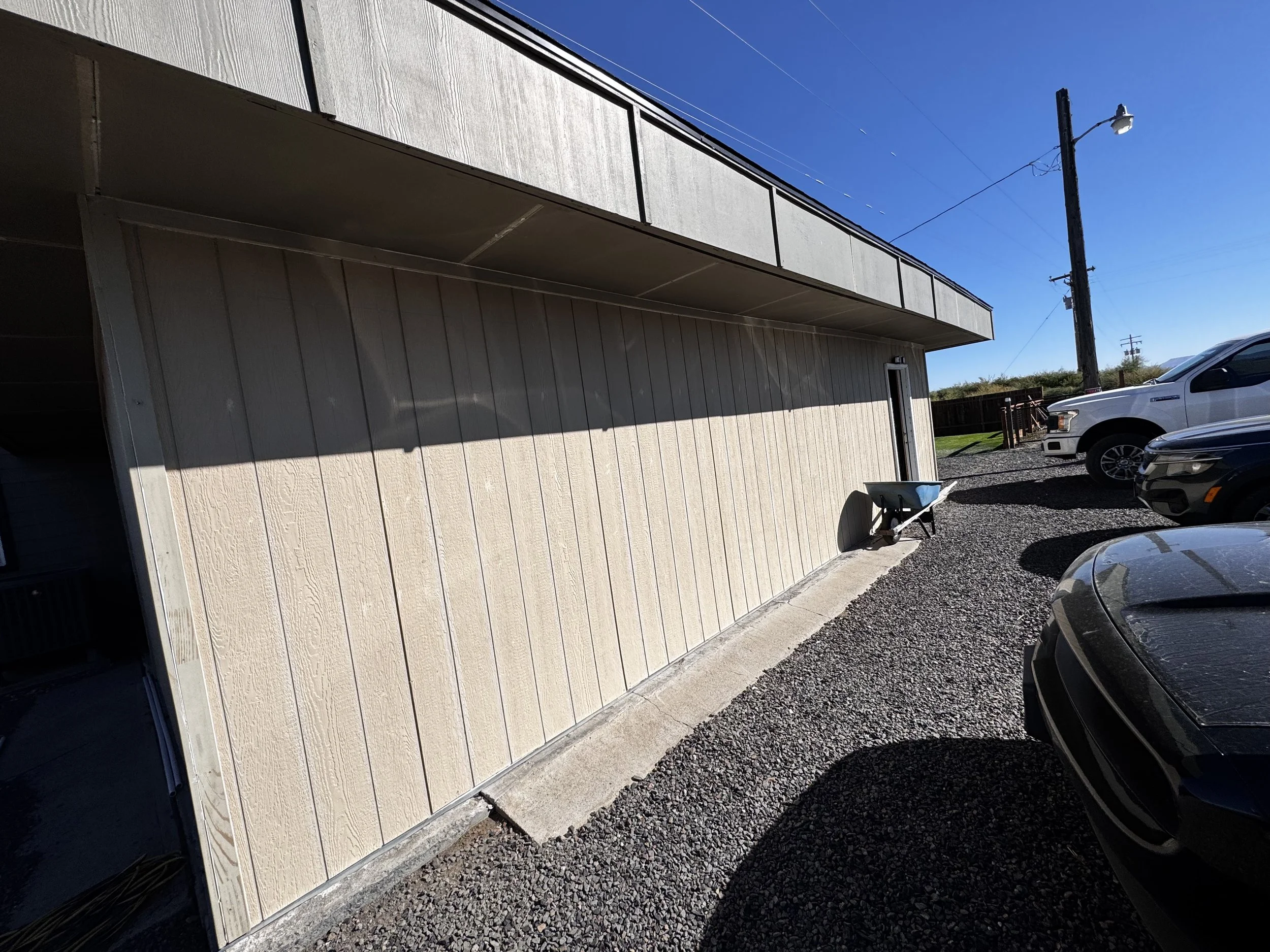 Side view of a beige building with a small door, parked white trucks, a wheelbarrow, and a utility pole under a clear blue sky.