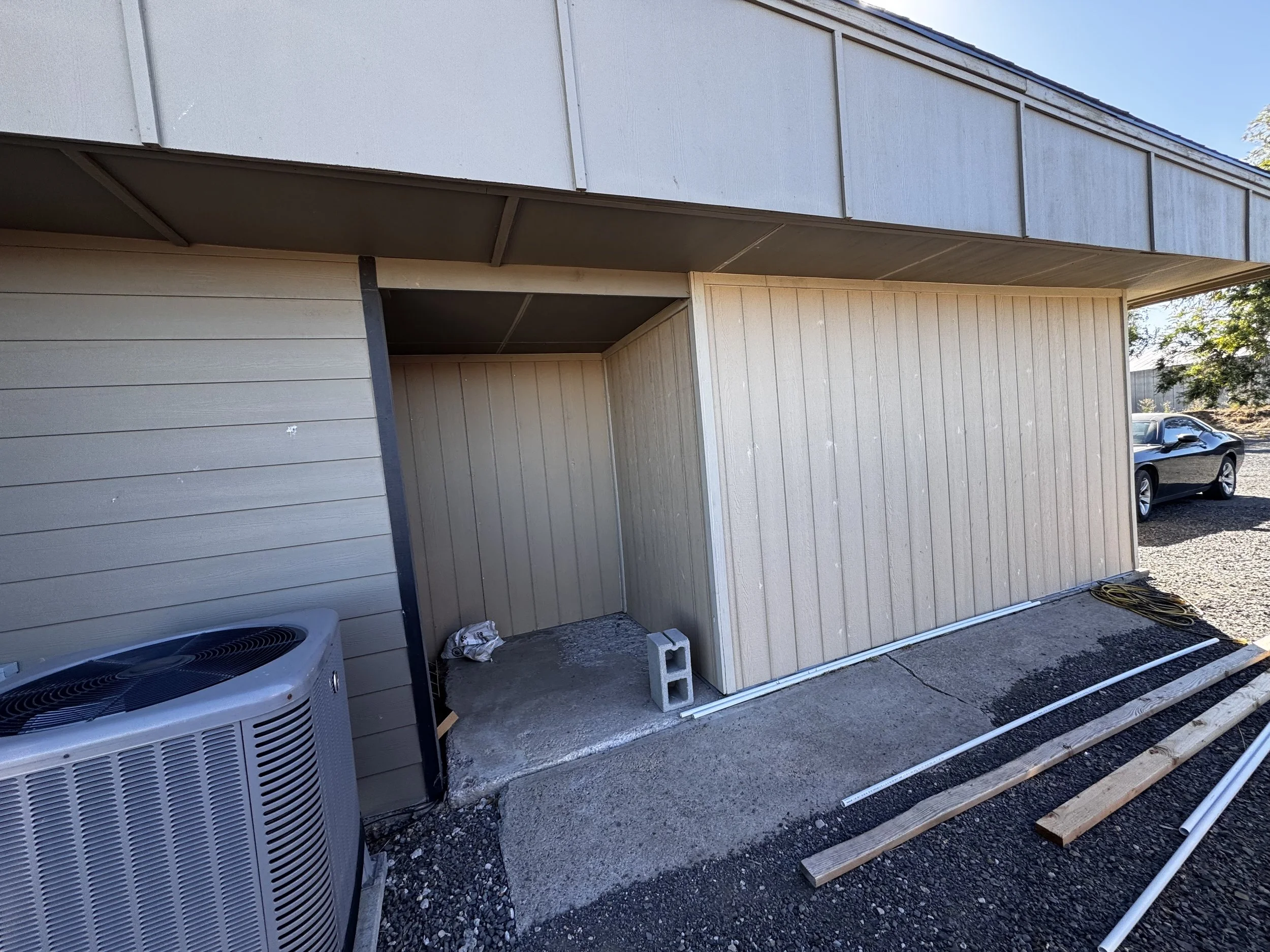 Construction site of a building with beige and gray siding. An air conditioning unit on the left, a gray concrete block in the middle, and building materials like metal pipes and wooden planks on the ground. The area appears to be under construction or renovation.