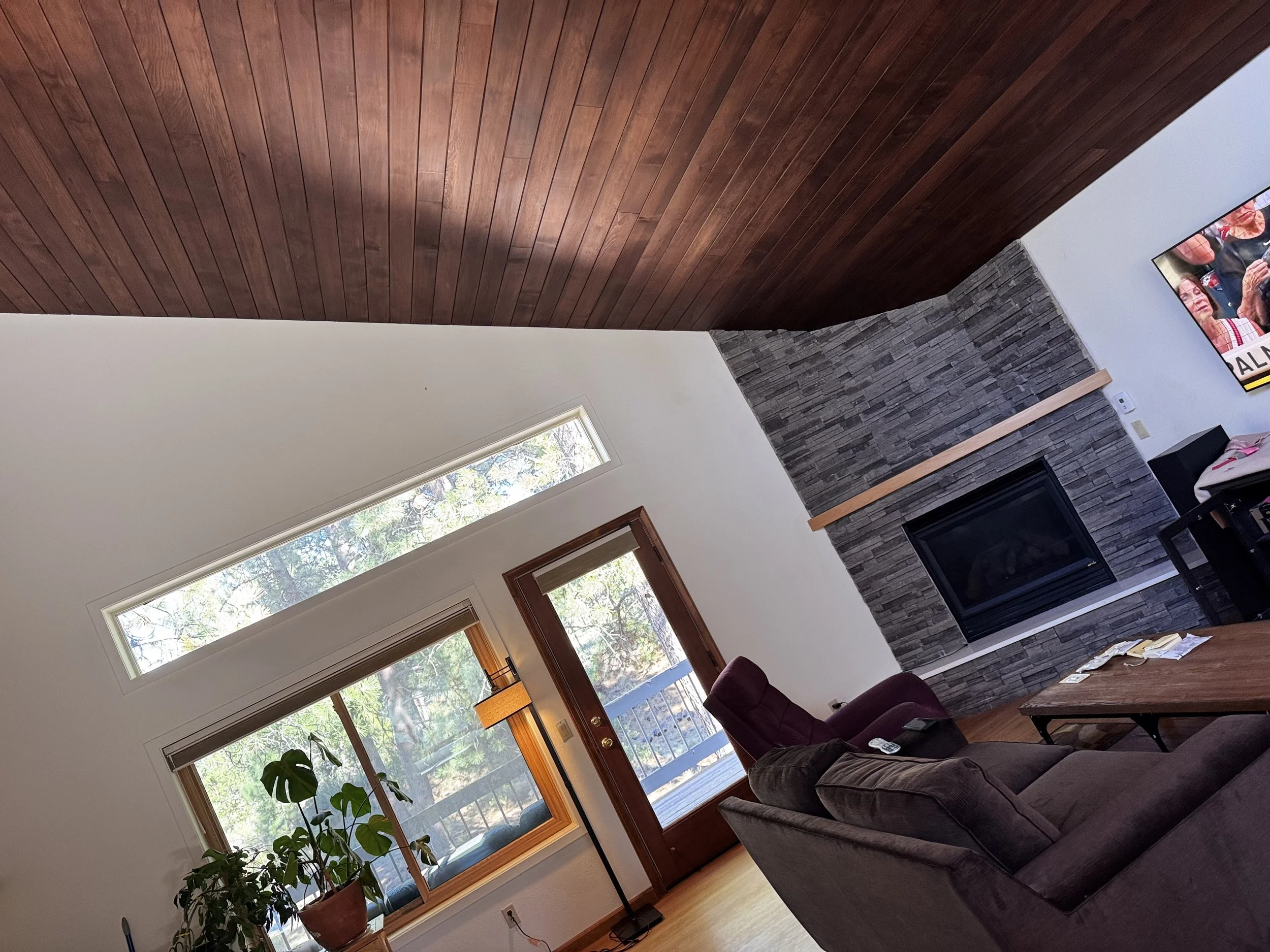 Living room with a stone fireplace, large windows, wooden ceiling, and brown furniture.
