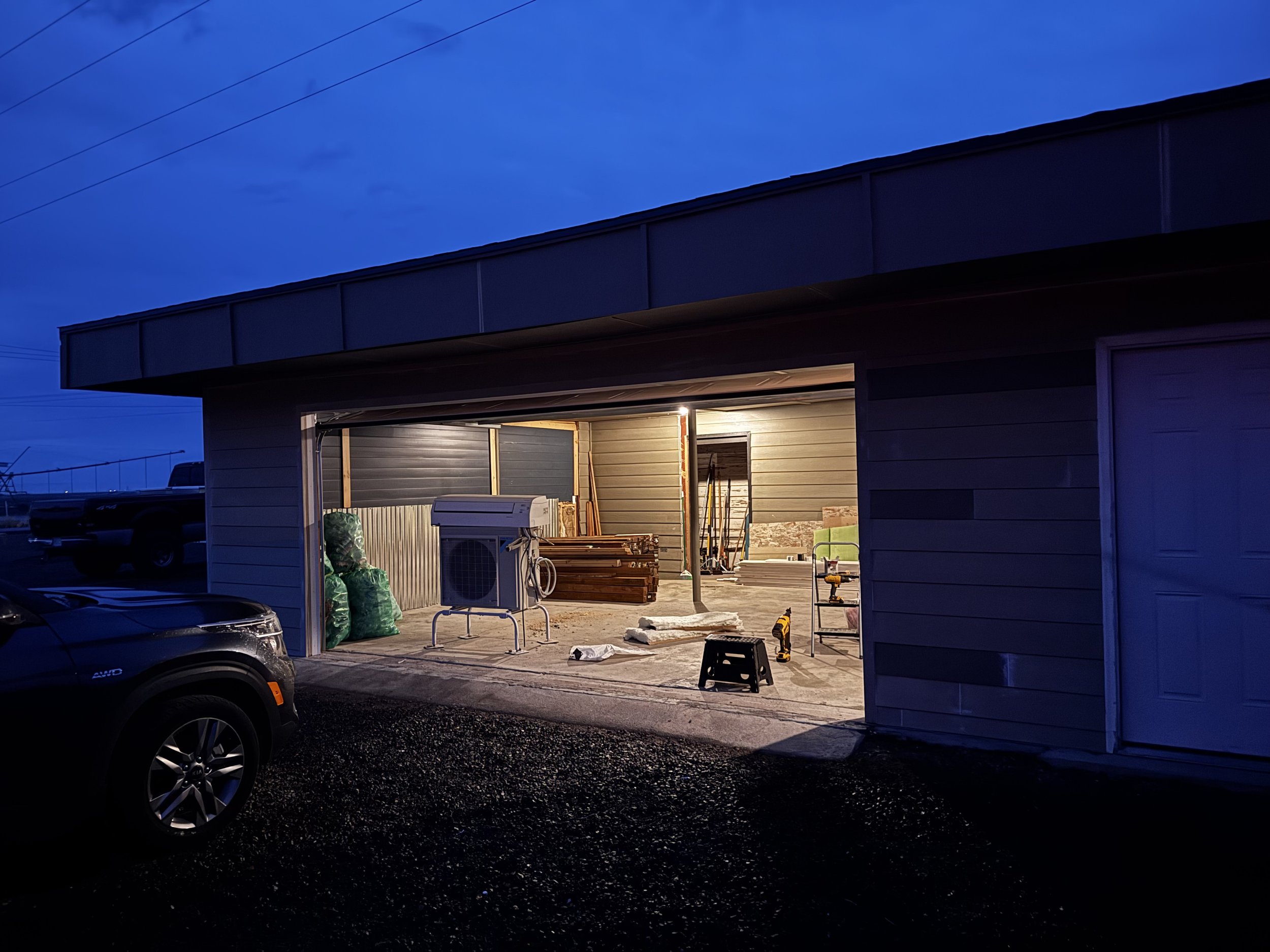 A garage with construction tools and materials inside, illuminated by bright lights, during dusk or night, with a parked vehicle outside.