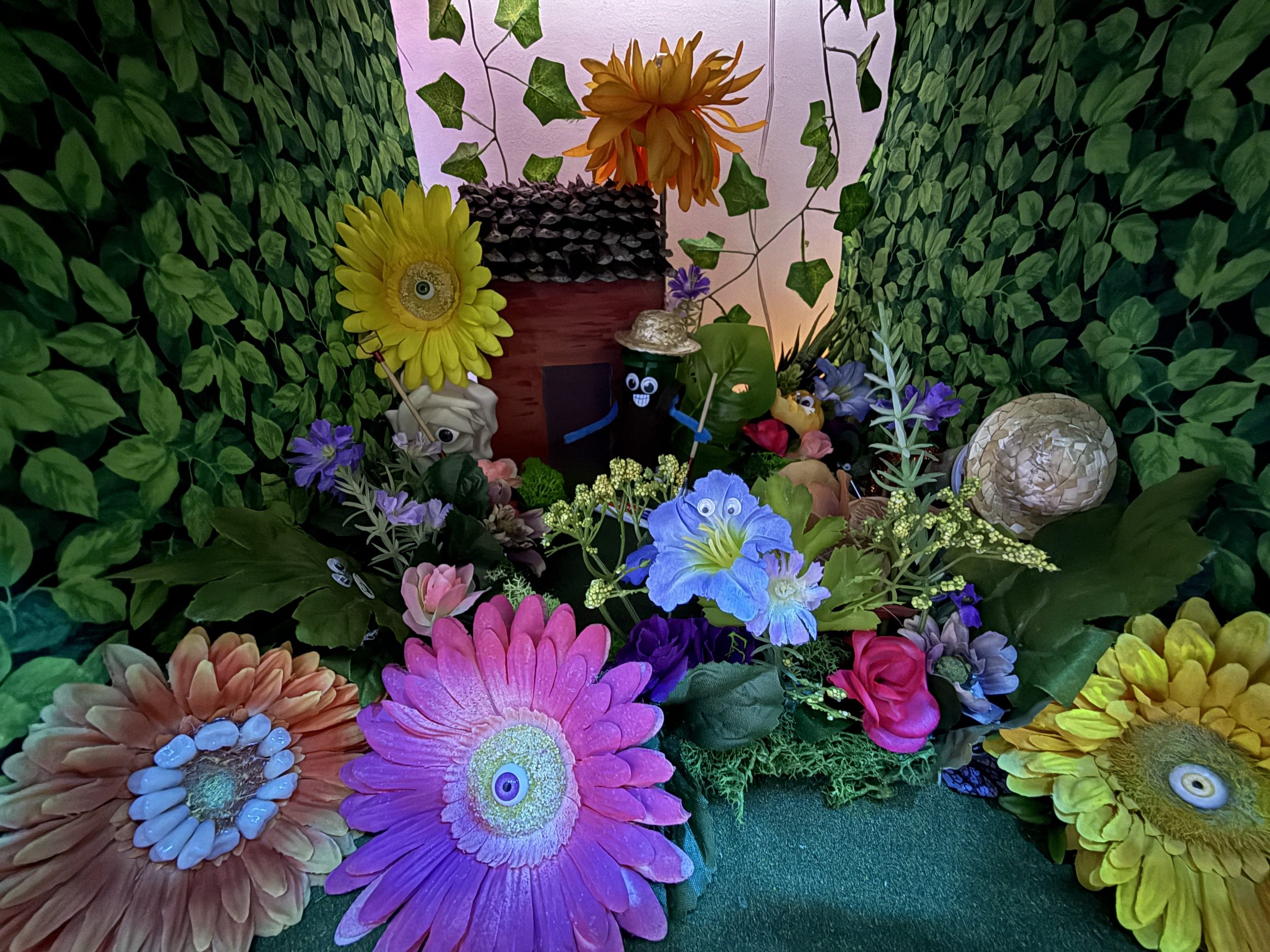 Eerie diorama of a floral cottage scene where all the flowers have eyes, and a pill bottle with eyes and limbs smiles at you.