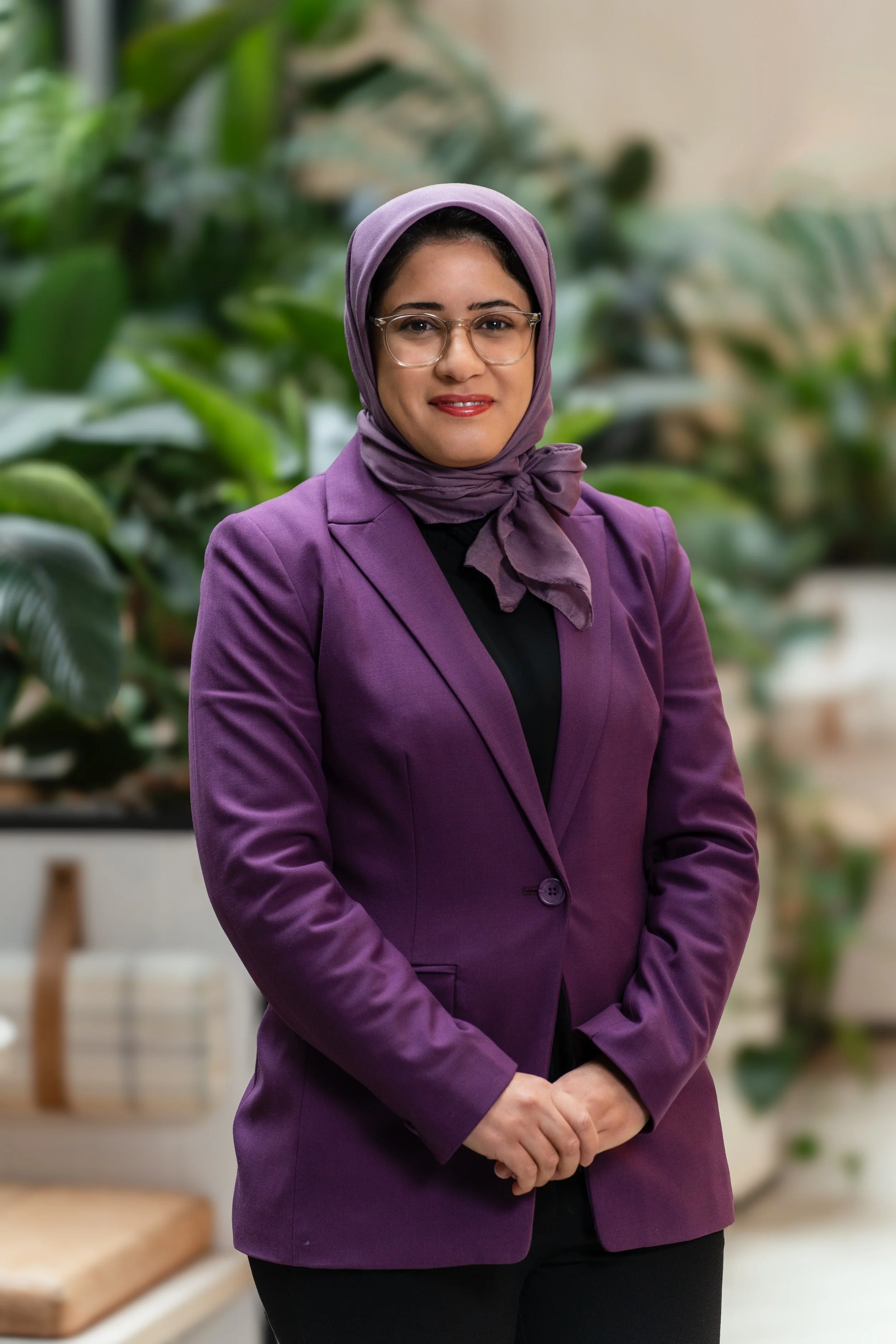 A woman wearing a purple blazer and hijab standing in a lush, green indoor garden space.