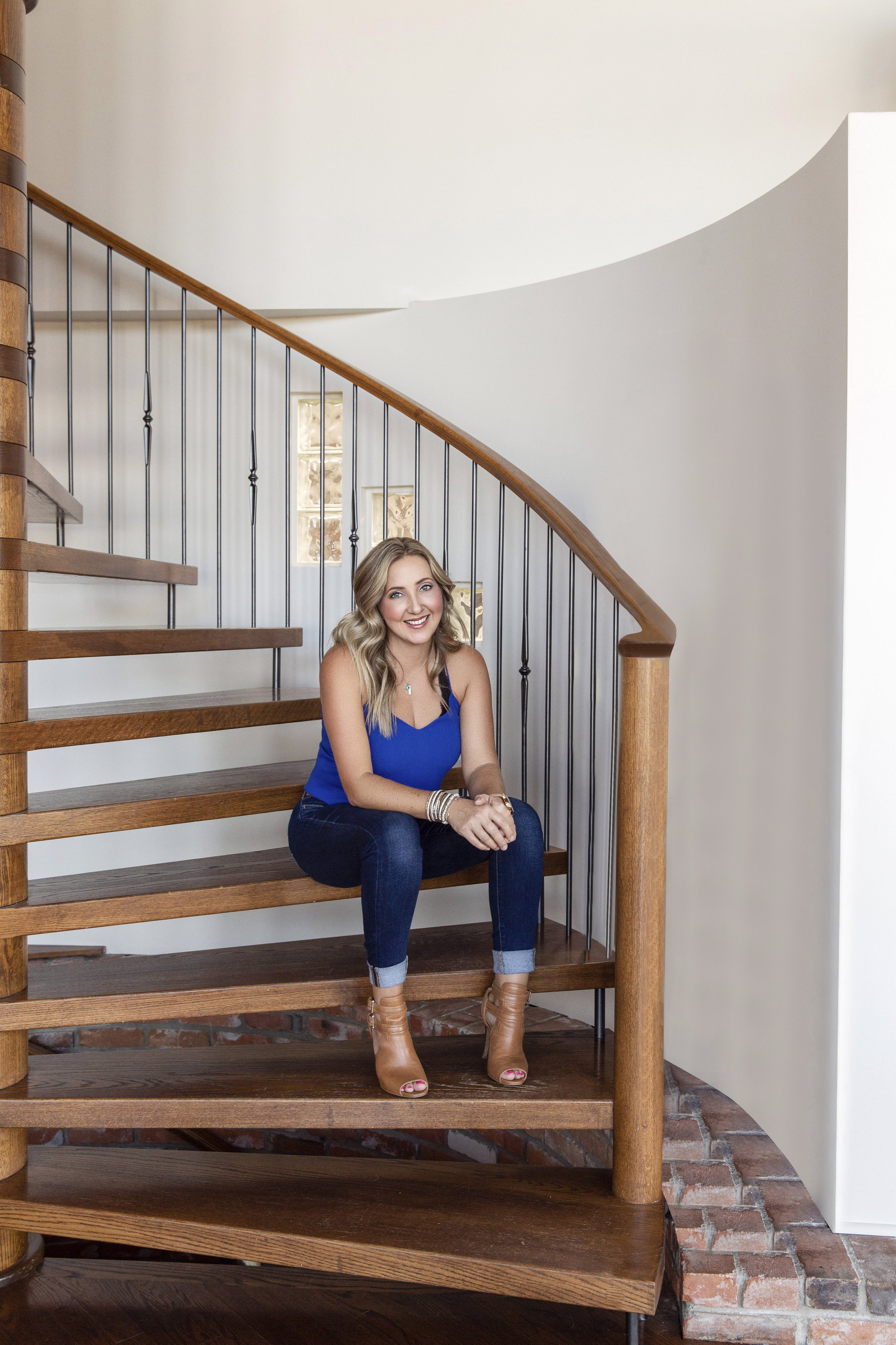 A woman smiling and sitting on a wooden spiral staircase, wearing a blue top, dark jeans, and tan ankle boots, with a white wall and small windows in the background.