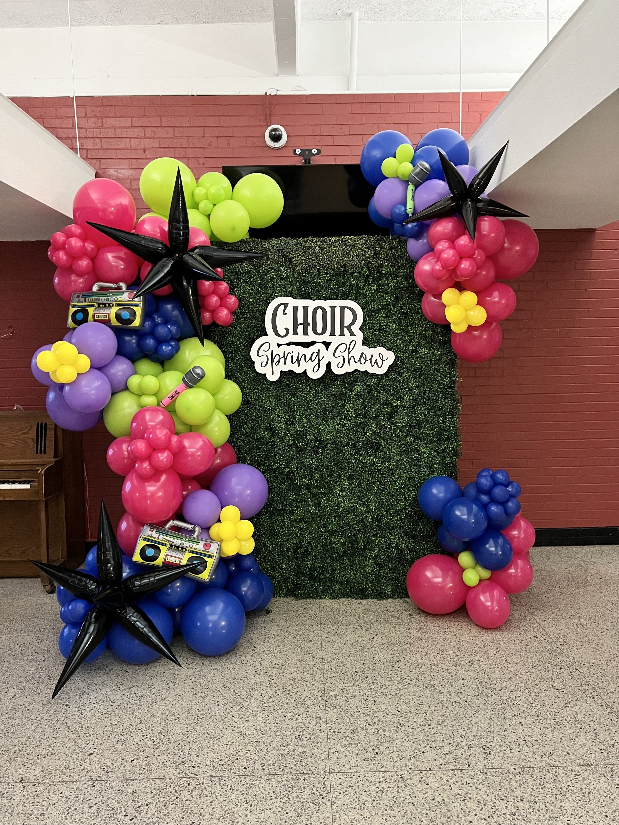 Decorative balloon arch with pink, purple, blue, green, and yellow balloons, featuring star-shaped black balloons, a miniature boombox, and a pink microphone, in front of a green hedge backdrop with a 