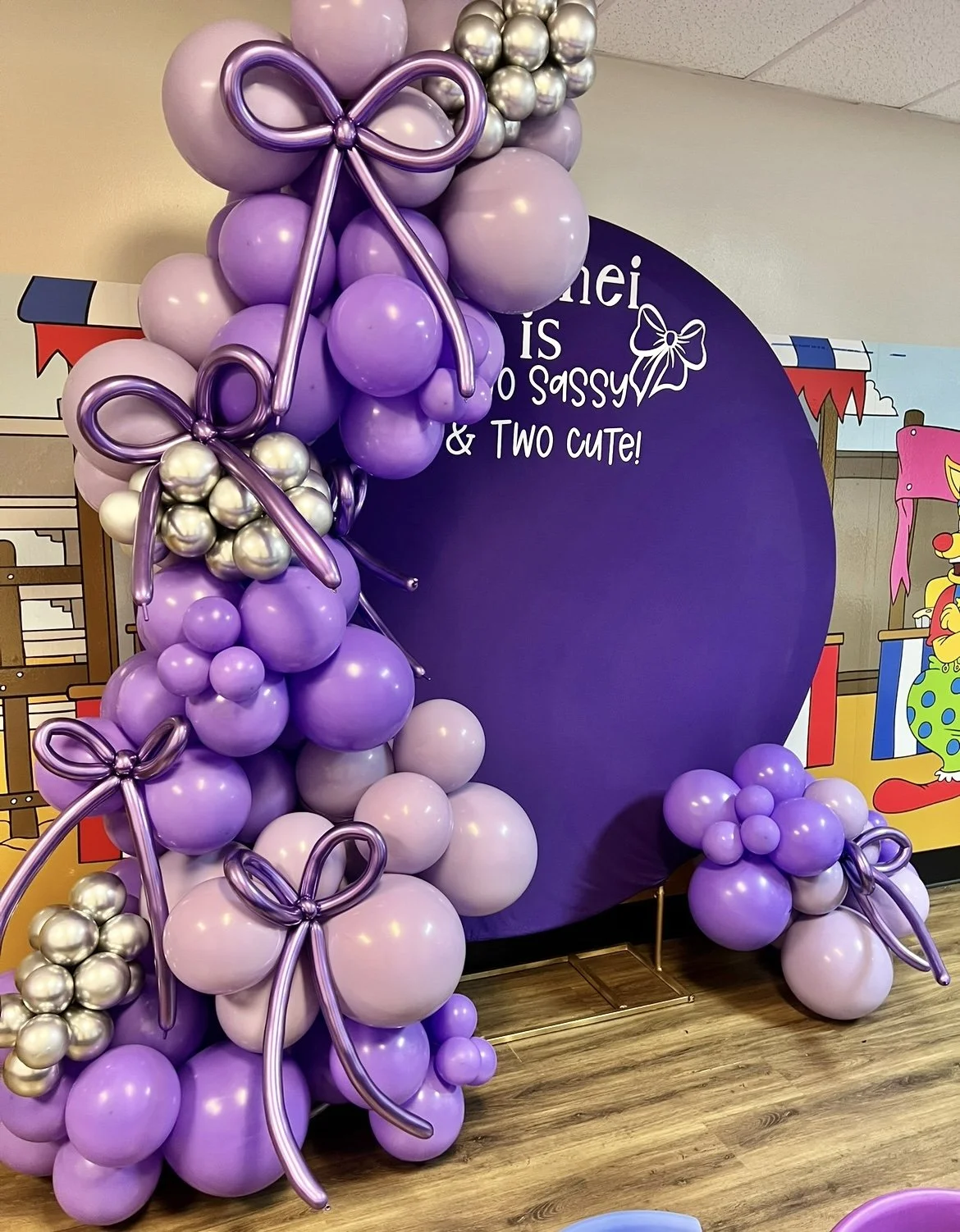 Purple and lavender balloon decoration with bows, set against a purple display board with partially visible text, in a colorful indoor setting.