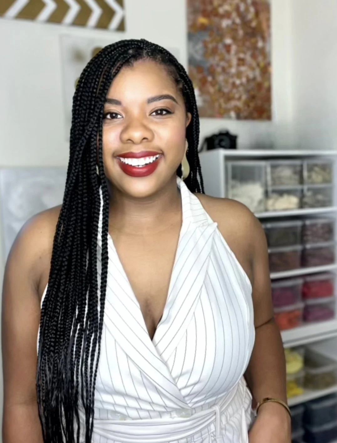 A smiling woman with long braided hair, wearing a sleeveless white striped top, standing in a room with colorful storage drawers and wall art in the background.