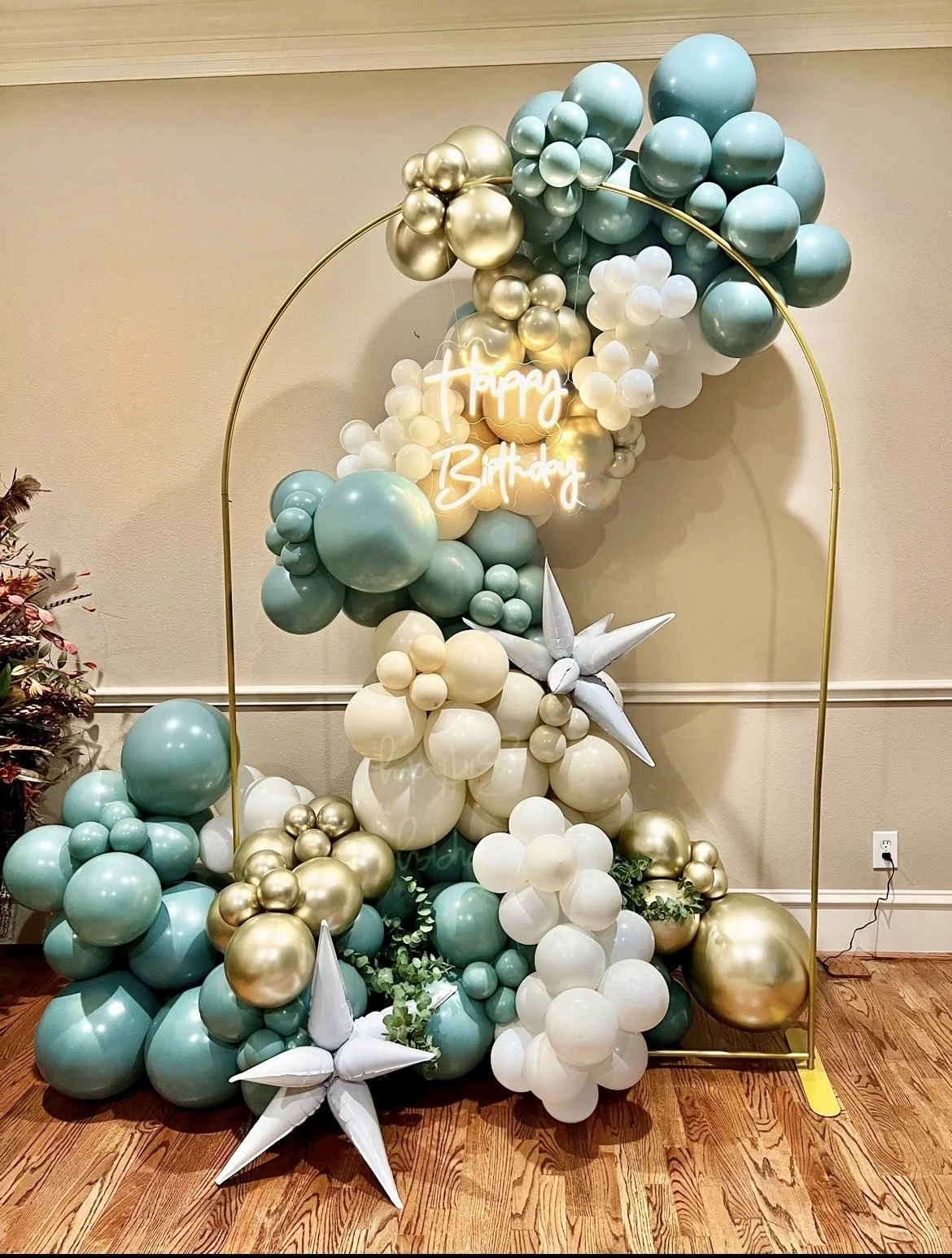 Decorative balloon arrangement with teal, white, and gold balloons, a neon 'Happy Birthday' sign, white star-shaped balloons, and greenery, framed by a gold arch, on a wooden floor.