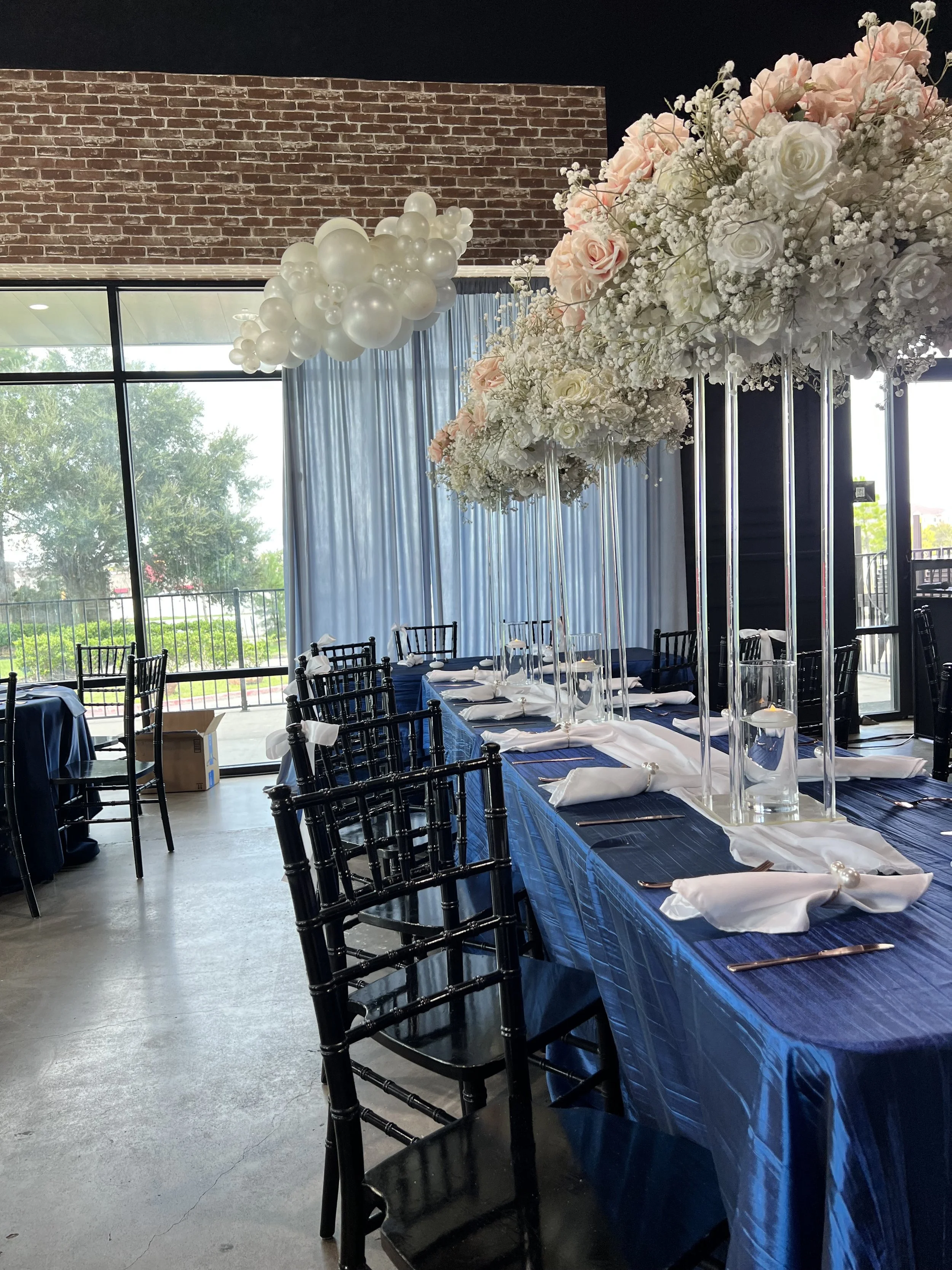 Elegant event table setup with black chairs, navy blue tablecloth, white napkins, tall floral centerpieces with white and blush pink roses, and glass candle holders inside tall glass cylinders, set in a room with large windows, blue curtains, and balloon decoration.