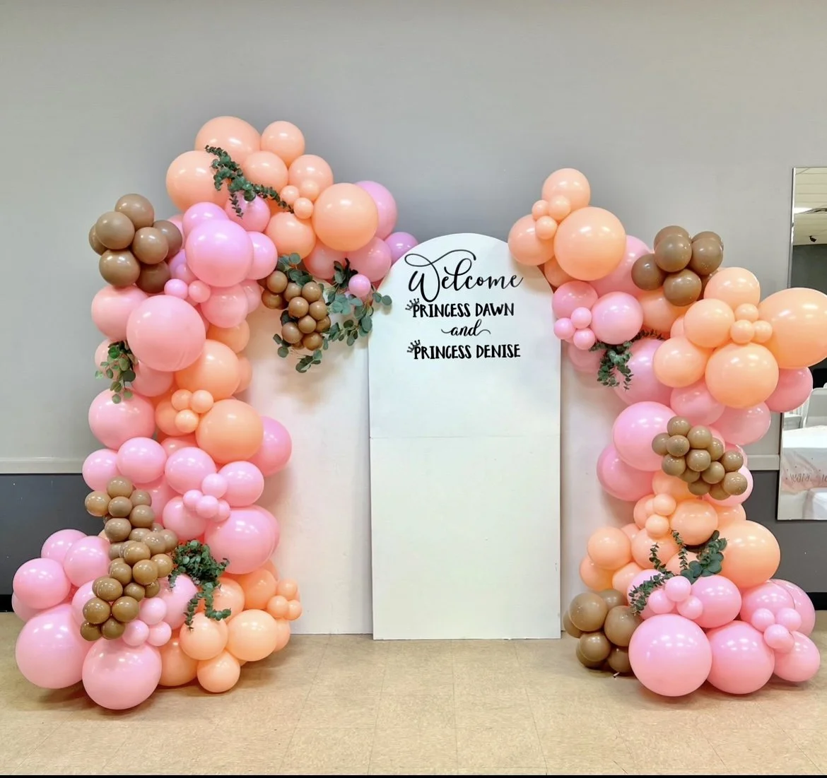 Balloon arch with pink and brown balloons surrounding a white sign that reads "Welcome Princess Dawn and Princess Denise".