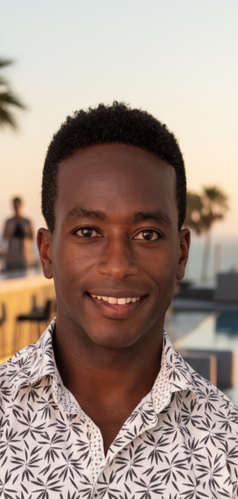 Young man smiling outdoors at sunset, wearing a patterned shirt, with palm trees and a body of water in the background.