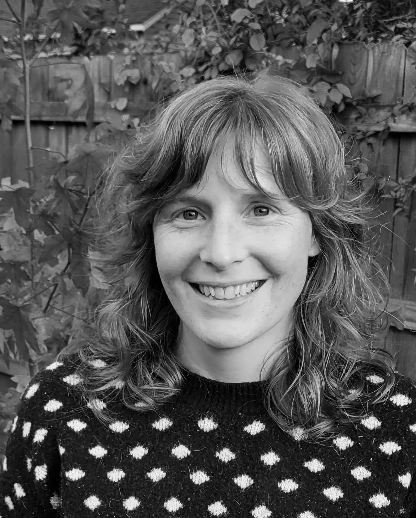 A woman with curly hair smiling outdoors, wearing a polka dot sweater, with a wooden fence and leafy plants in the background.