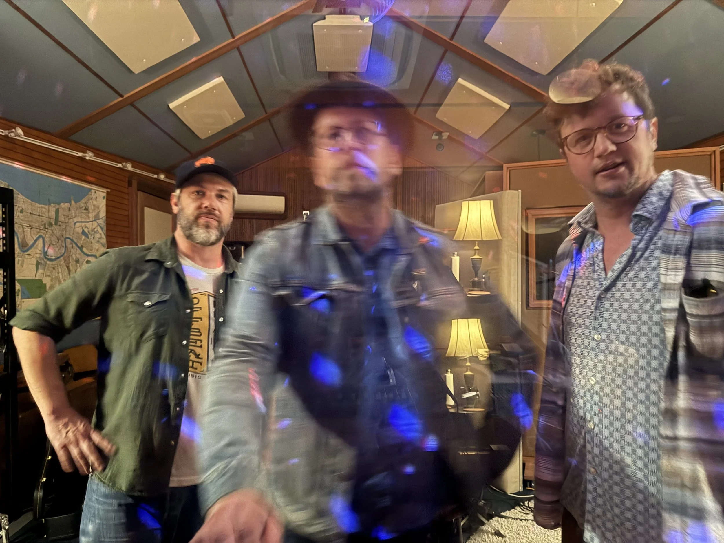 Three men in a music studio with wood-paneled walls and acoustic panels, illuminated by yellow lamps and colorful lighting, with a blurry effect in the center.