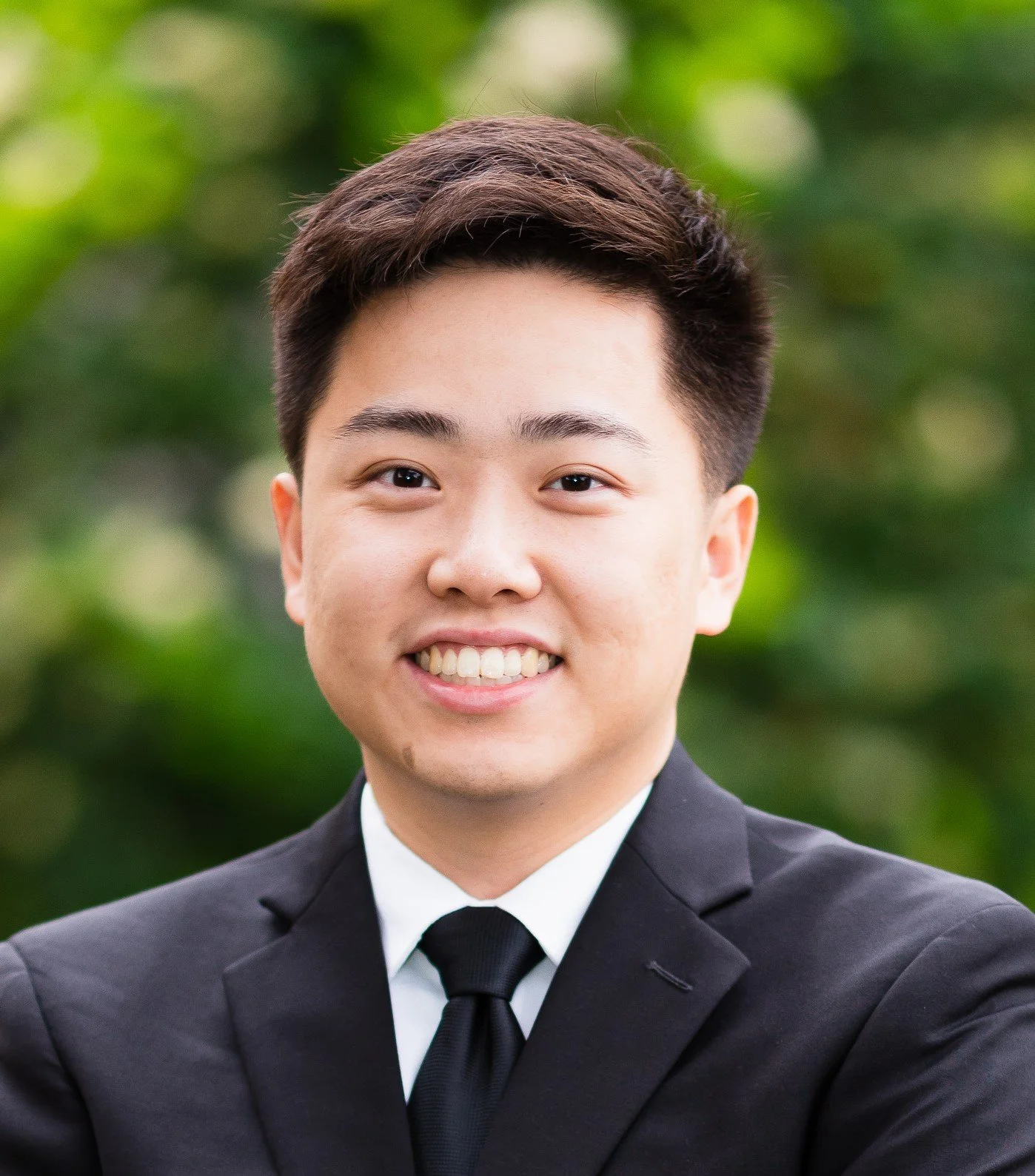 A young man in a black suit, white shirt, and black tie smiling outdoors with green blurred foliage in the background.