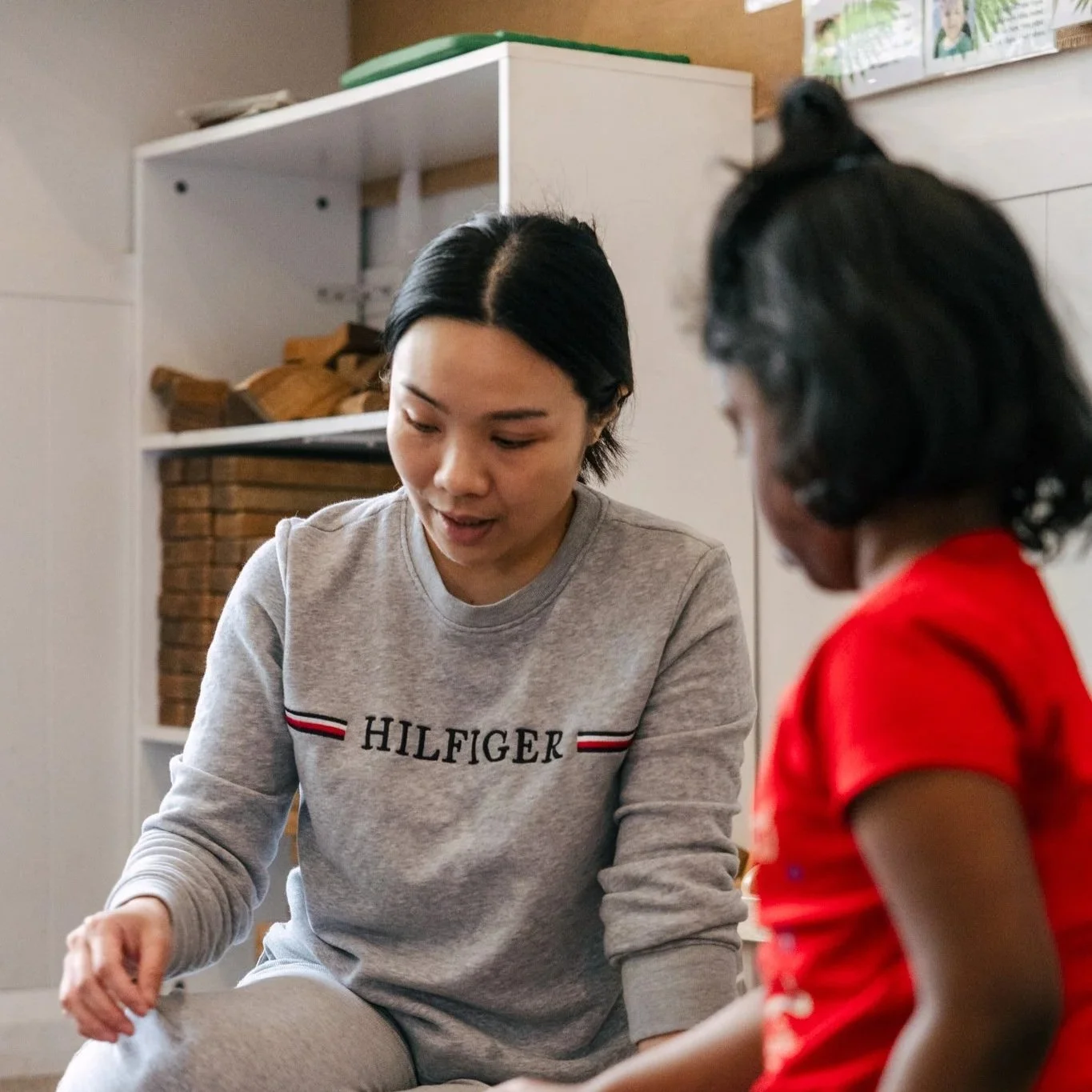A woman and a young girl sitting on the floor, engaged in an activity together. The woman is wearing a gray Tommy Hilfiger sweatshirt, and the girl is wearing a red shirt. They appear to be focused on something in front of them.